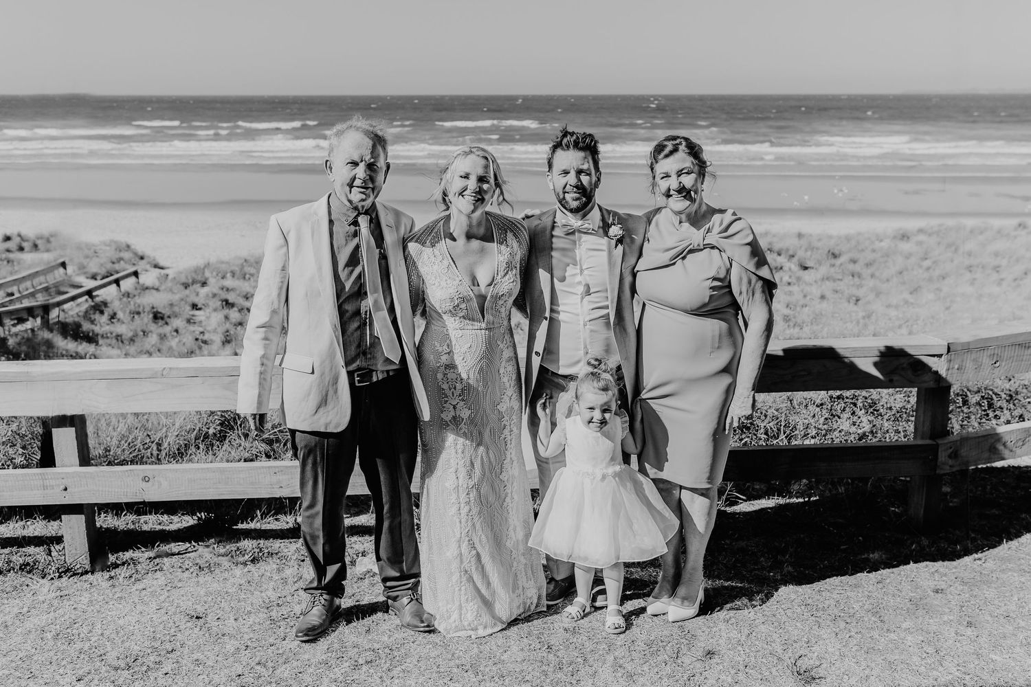 A small wedding group poses together on a coastal overlook with the ocean visible behind them. Papamoa Surf Lifesaving Club Wedding