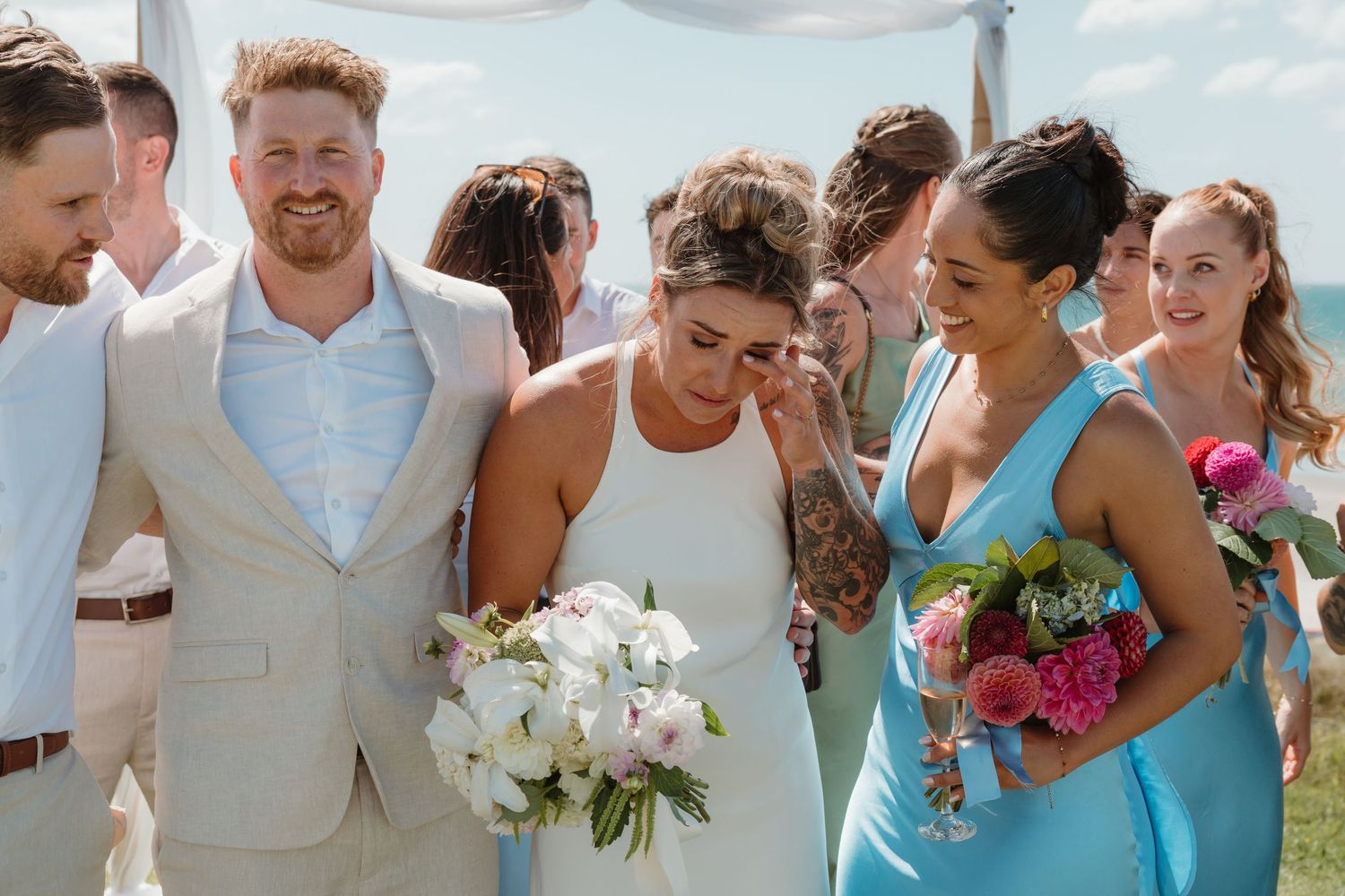 Bride and groom walking down the aisle with bridesmaids in blue dresses holding colorful bouquets at outdoor wedding.