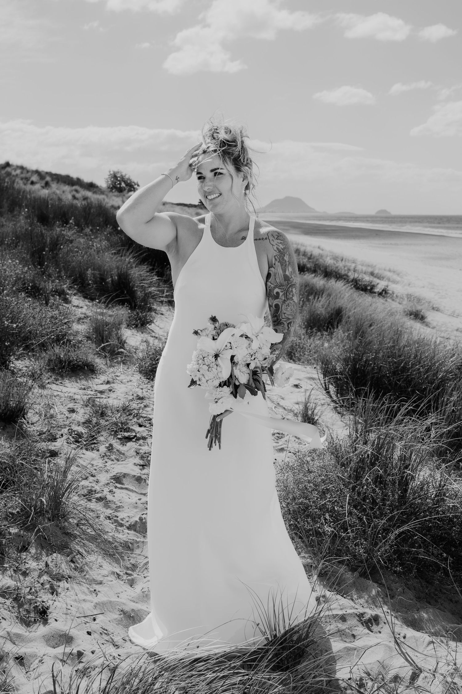Bride in white halter gown holds bouquet on sandy beach dunes, black and white wedding photography.