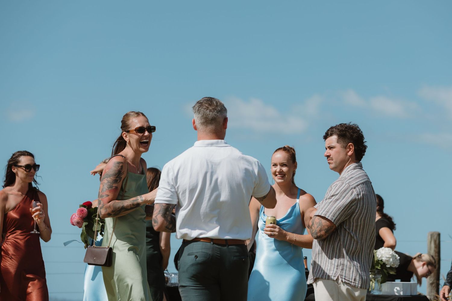 Guests socializing outdoors at a sunny wedding reception, with a tattooed woman in mint dress and man in white shirt.