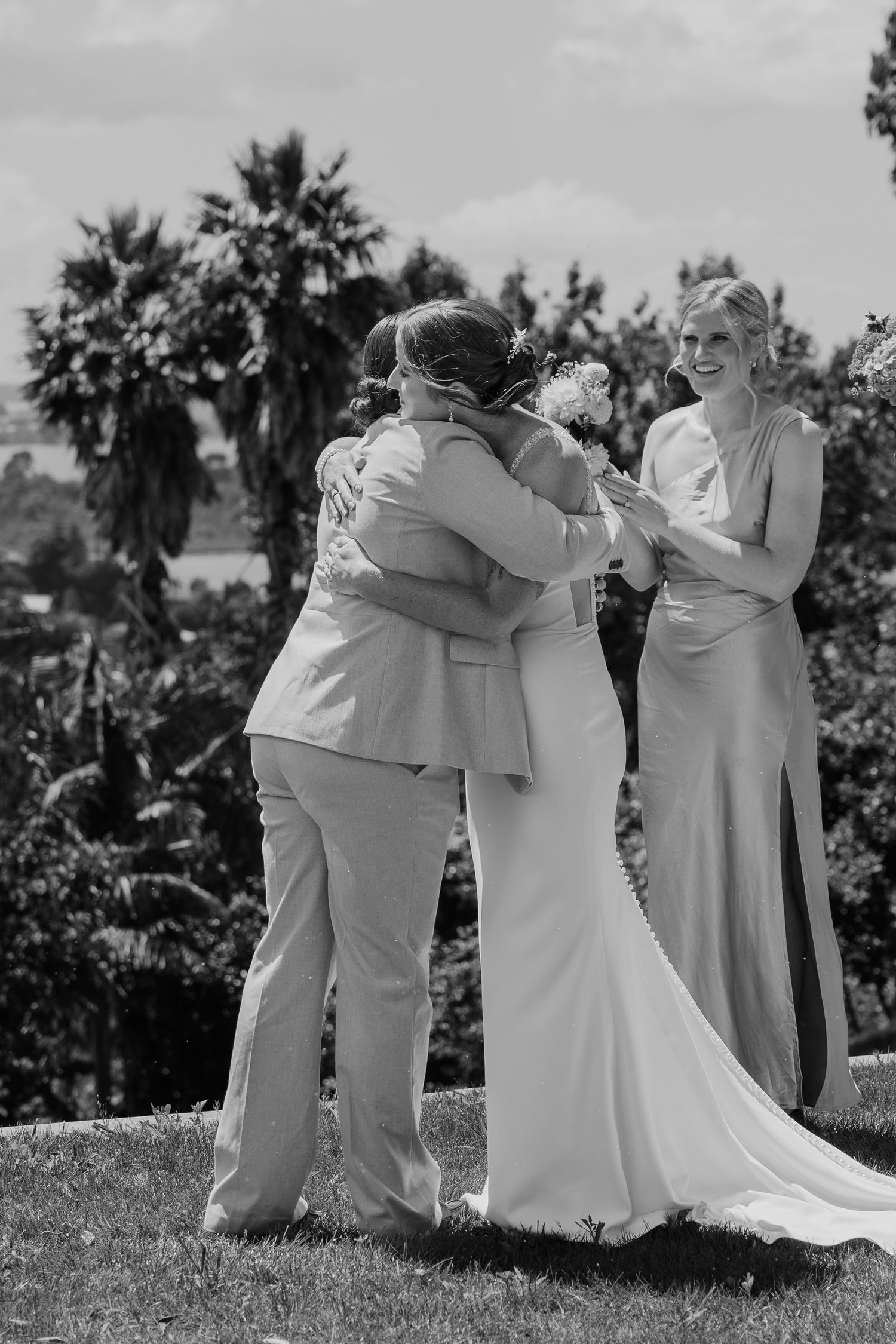 Black and white photo of two brides embracing at outdoor wedding ceremony with bridesmaid holding bouquet.
