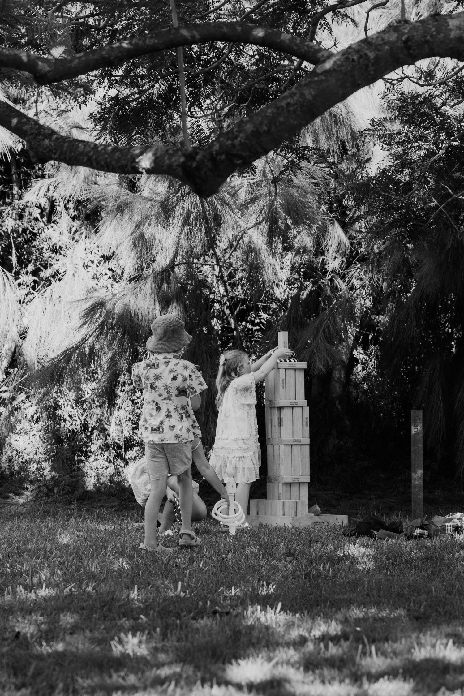 Children playing with wooden blocks outdoors under large trees in a black and white photo.