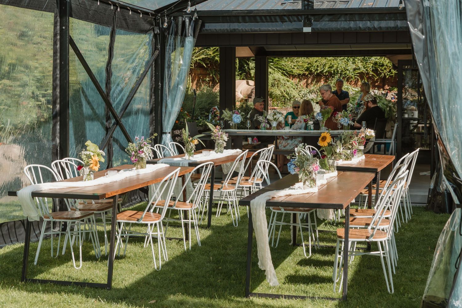 Outdoor wedding reception setup with wooden tables, white metal chairs, and floral centerpieces under a clear tent on green grass.