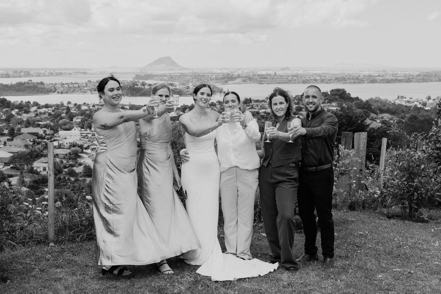 Black and white photo of bride with bridesmaids and guests celebrating outdoors with scenic coastal views.