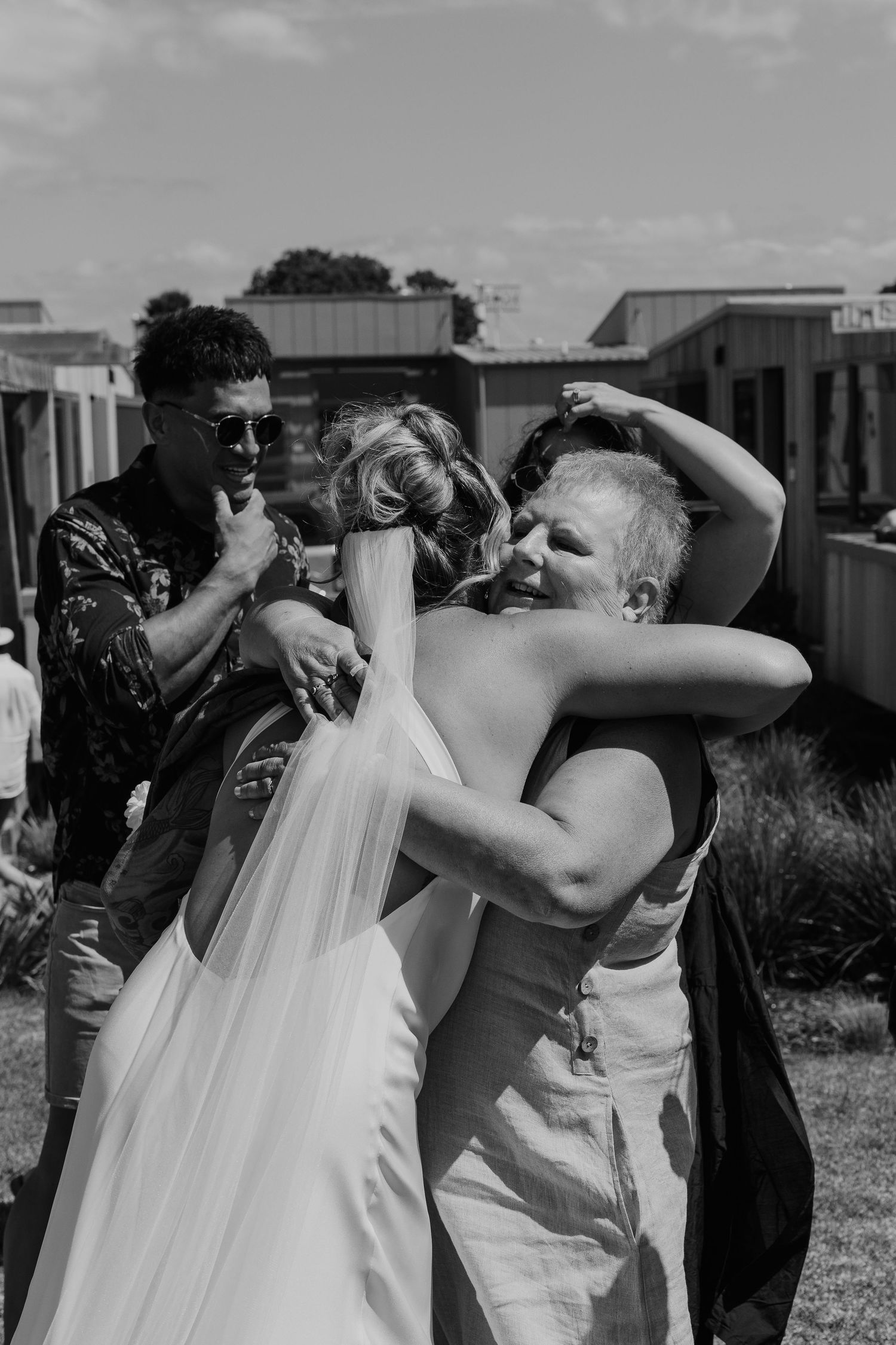 Black and white photo of a bride in a wedding gown and veil sharing a warm hug with an older woman at an outdoor wedding.