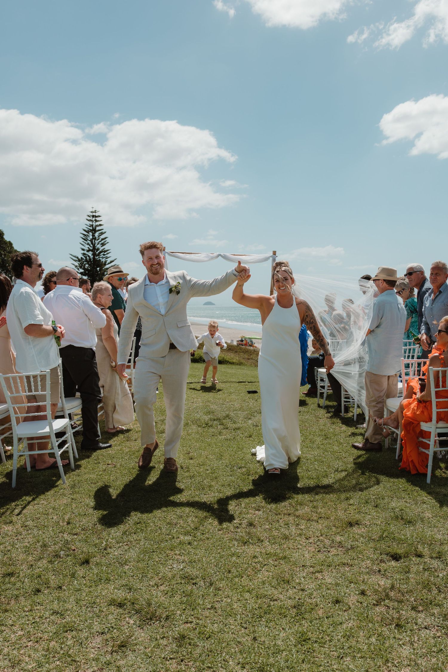 Joyful newlyweds celebrate walking back down the aisle at an outdoor beach wedding ceremony with guests cheering.