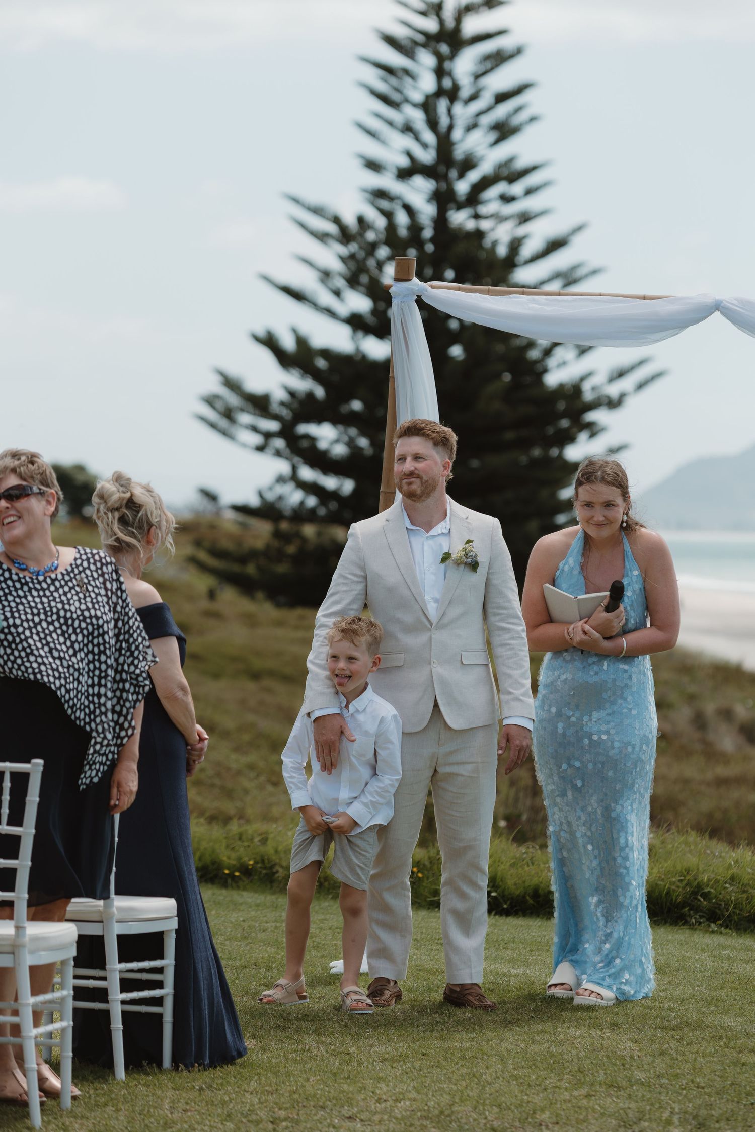 Groom in beige suit walks with young boy and bridesmaid in blue dress at outdoor beach wedding ceremony.