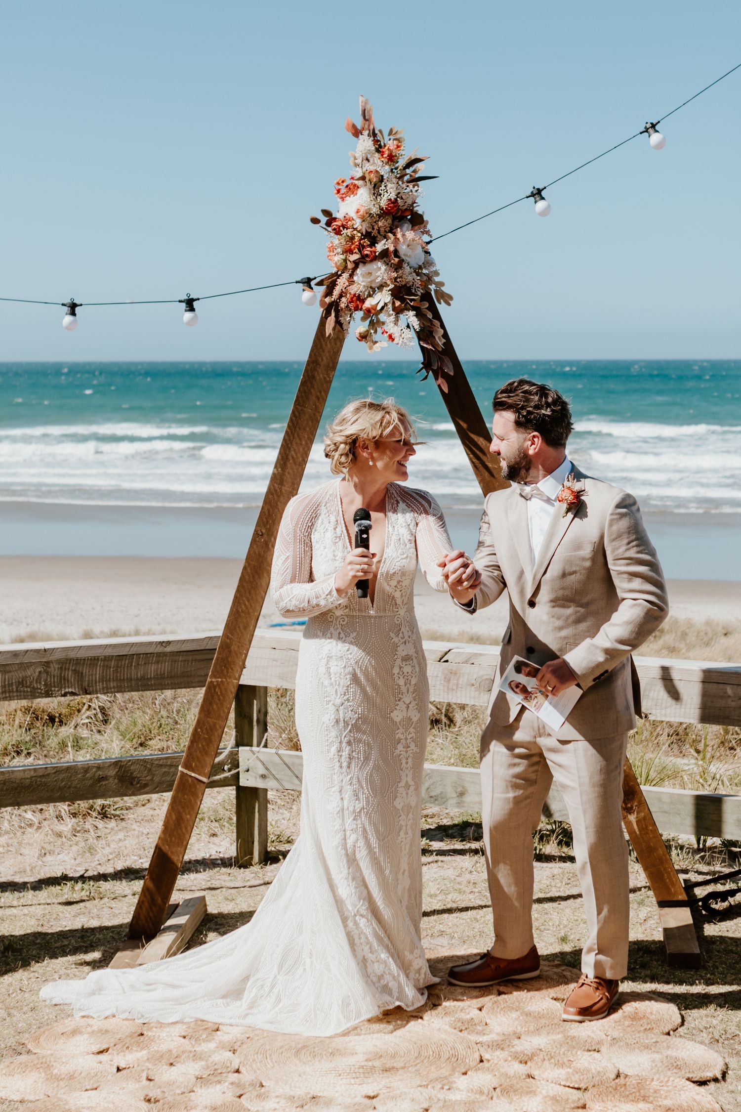 A beach wedding ceremony setup with wooden arch, string lights and floral decorations along the ocean shore.