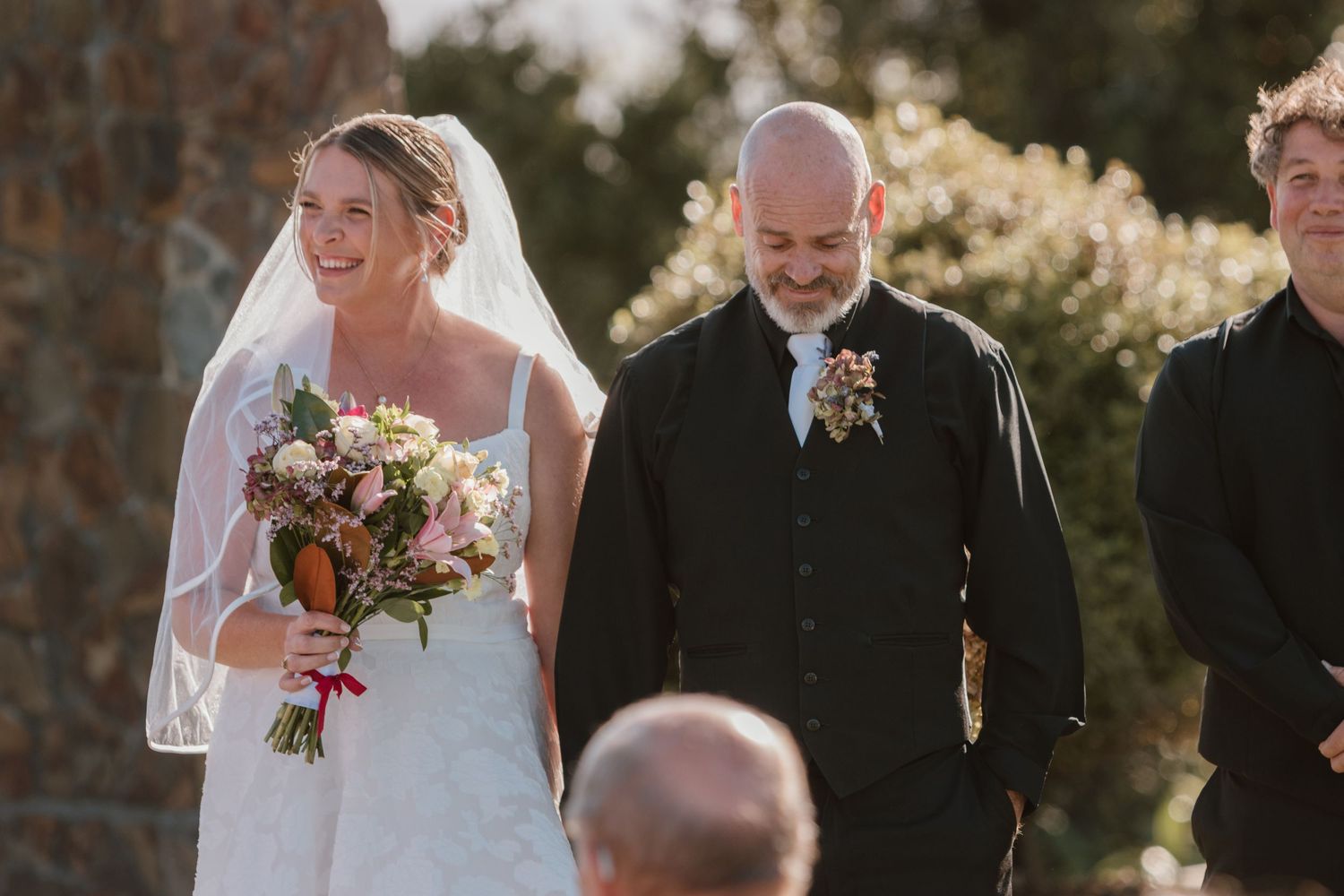 An outdoor wedding ceremony in warm sunlight featuring a couple and officiant with colorful floral bouquet.