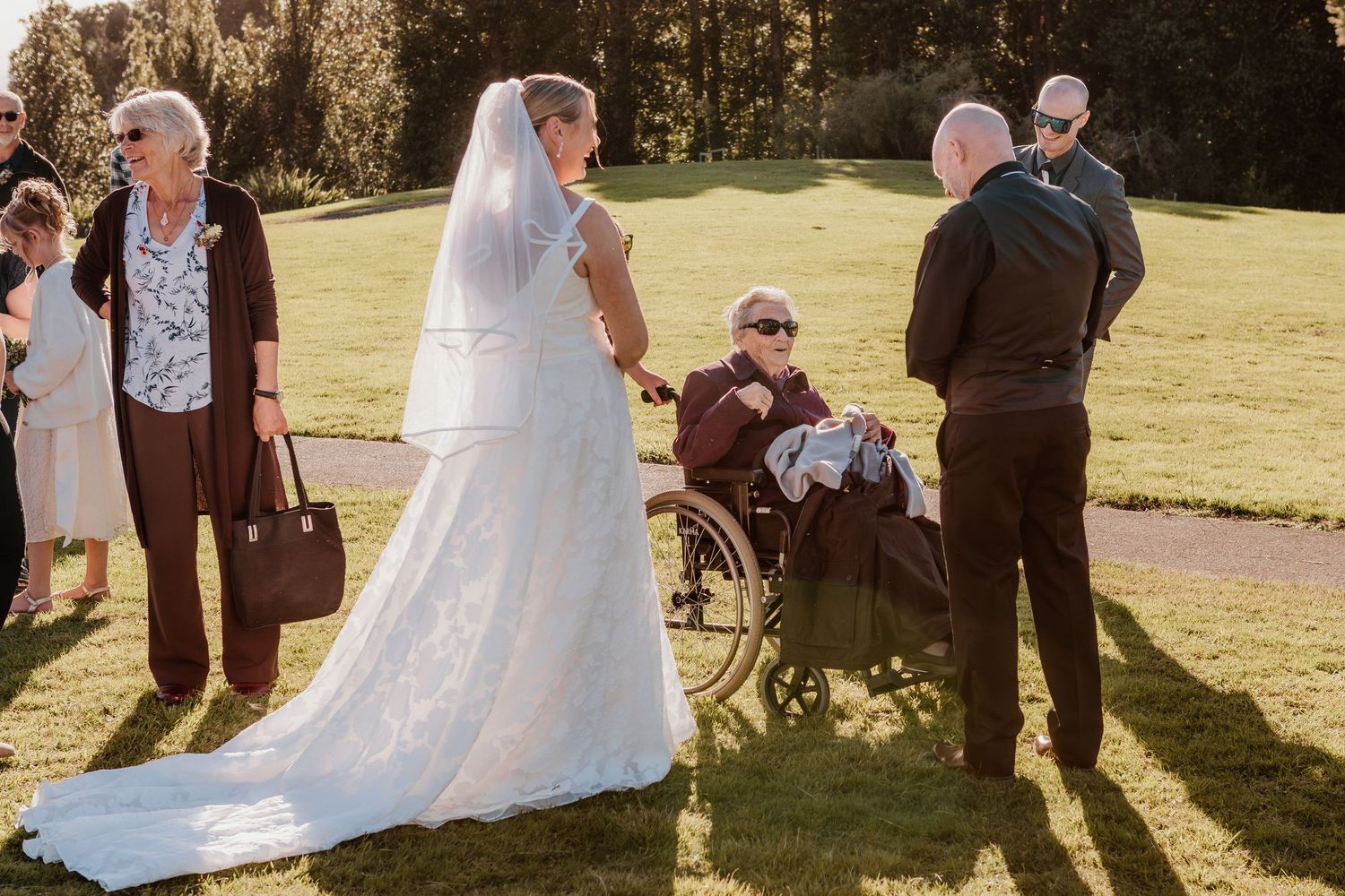 A rustic outdoor wedding ceremony in a sunlit garden setting with white decor and guests seated on wooden chairs.