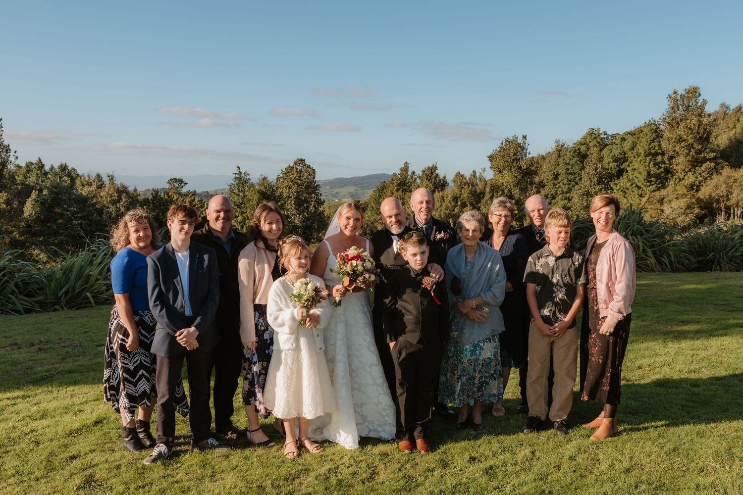Wedding party gathers outdoors on green lawn at sunset with mountain landscapes in background.