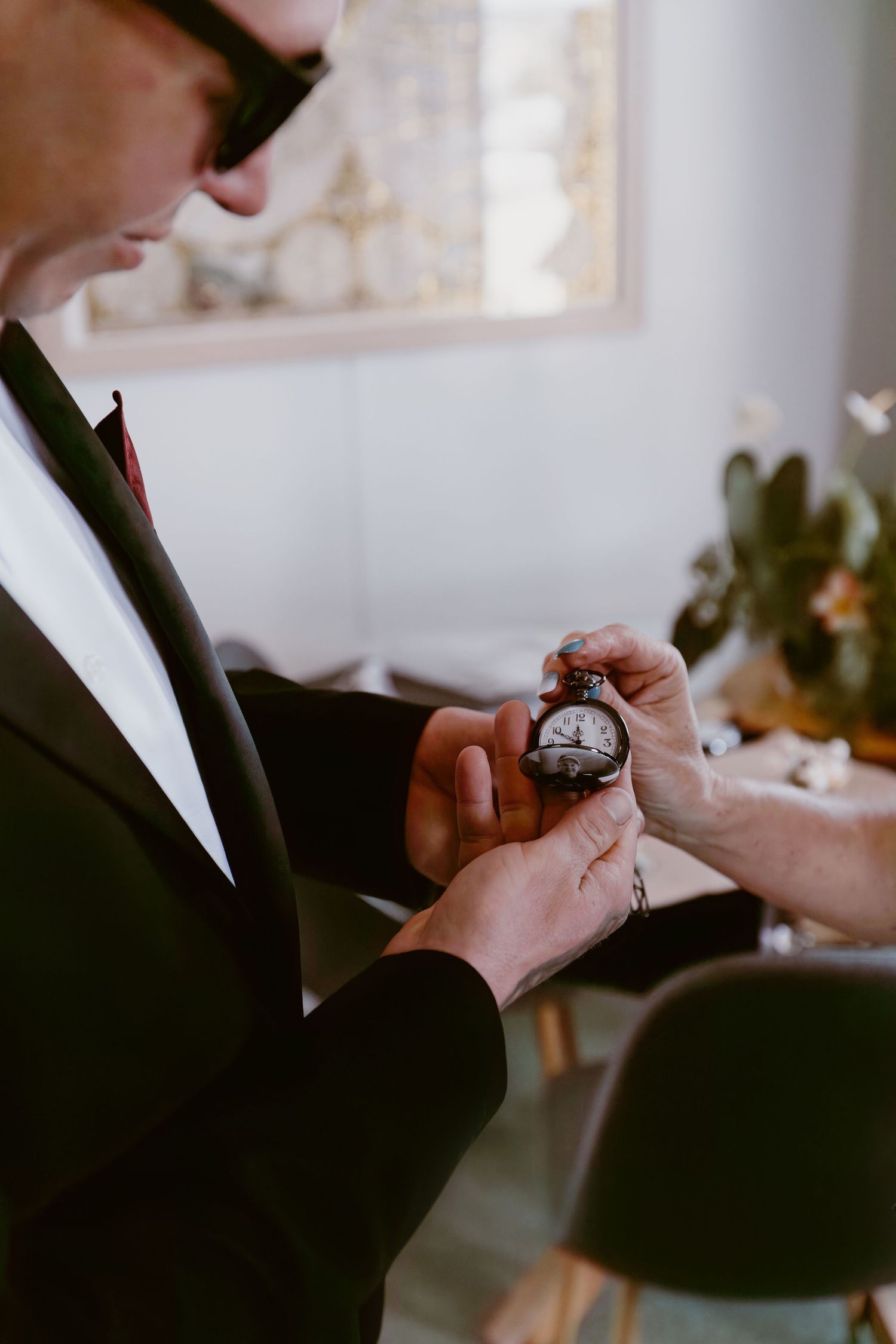 Person in formal black suit holding and examining a vintage pocket watch in decorative indoor setting.