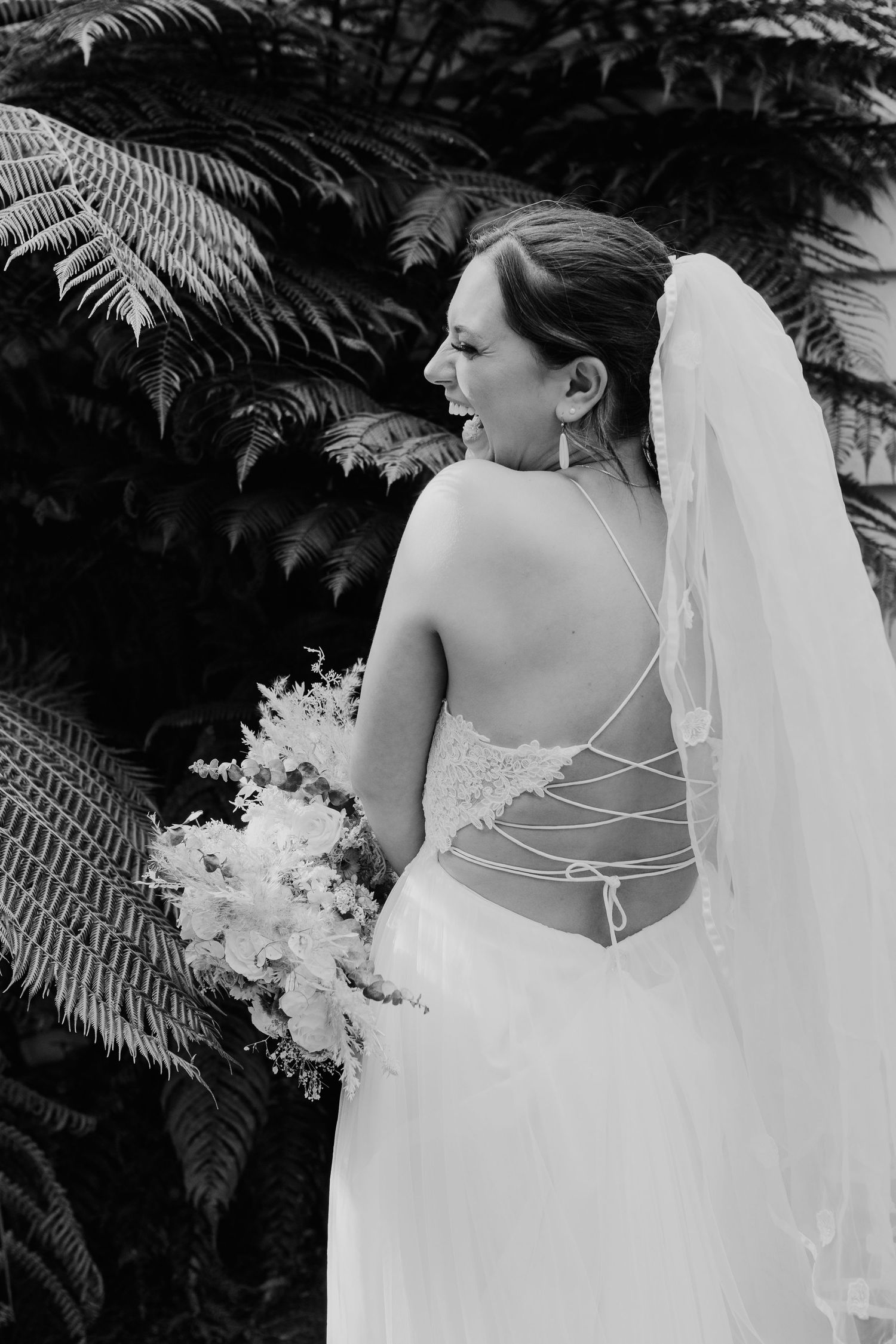 Black and white wedding photography series showing bride's detailed back lace dress against tropical fern backdrop.