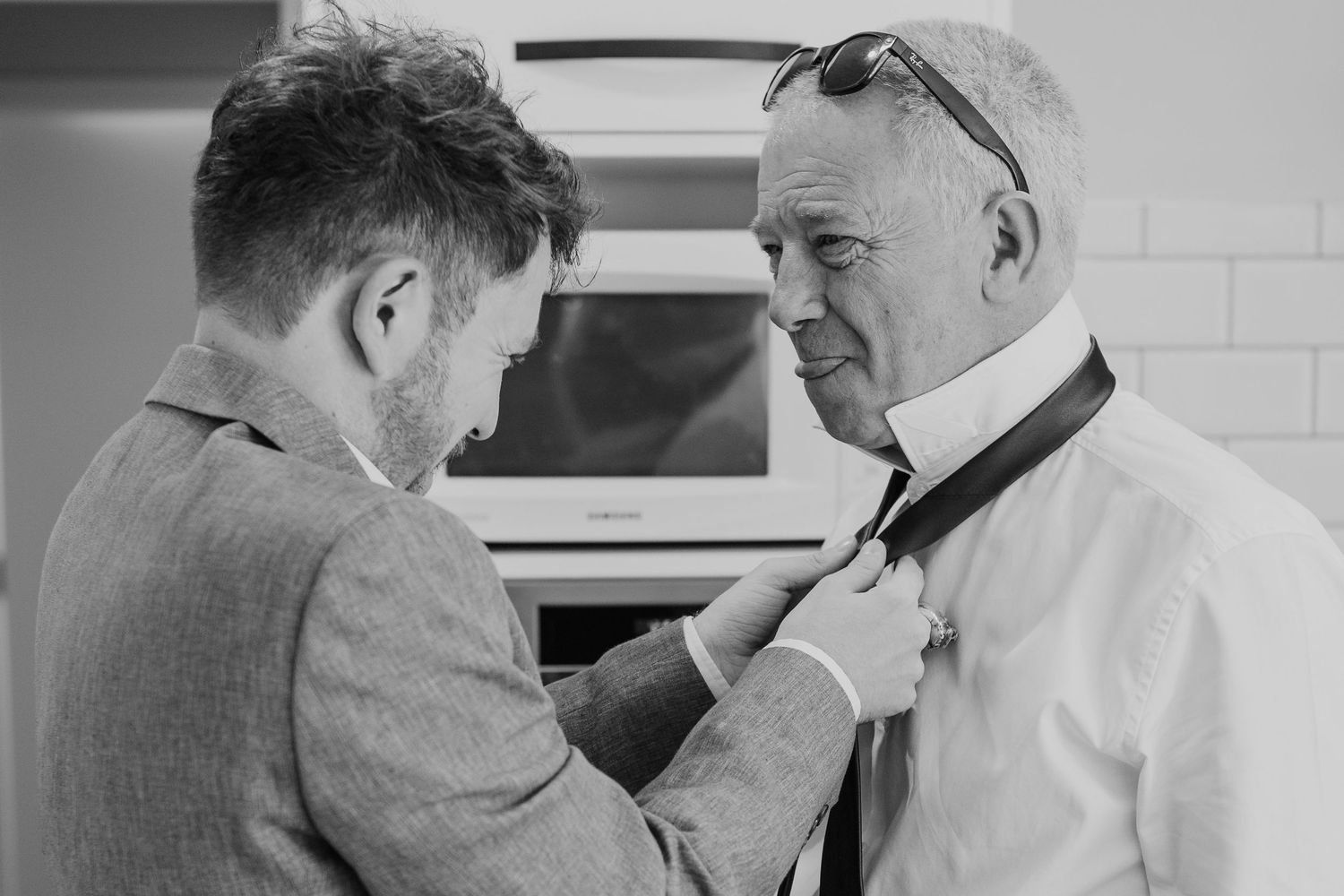 A candid black and white photograph shows two men in conversation, with one adjusting the other's necktie.