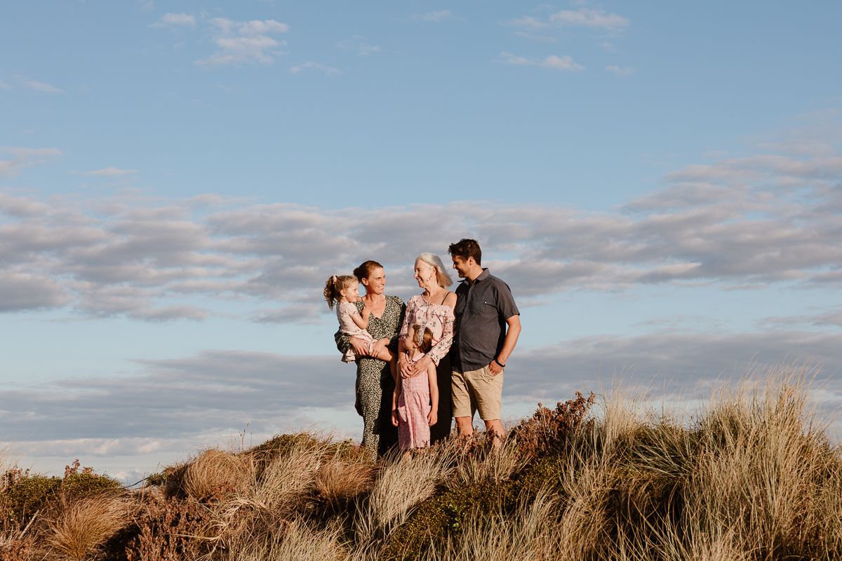People standing on a hilltop with tall grass against a blue cloudy sky during golden hour. Tauranga extended family photography with grandma