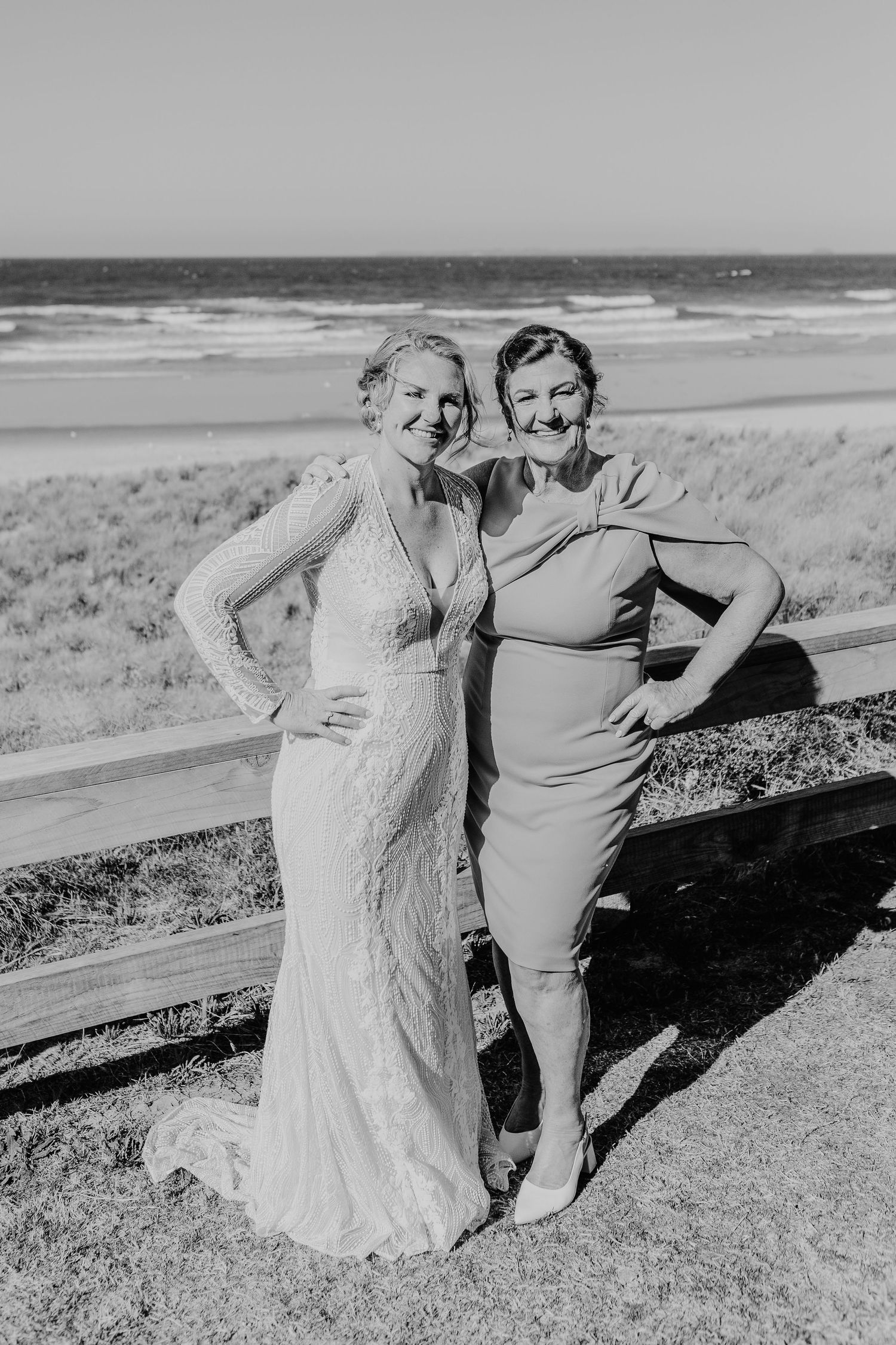 Two figures pose together on a coastal overlook wearing formal attire with ocean waves visible in the distance. Papamoa Surf Lifesaving Club