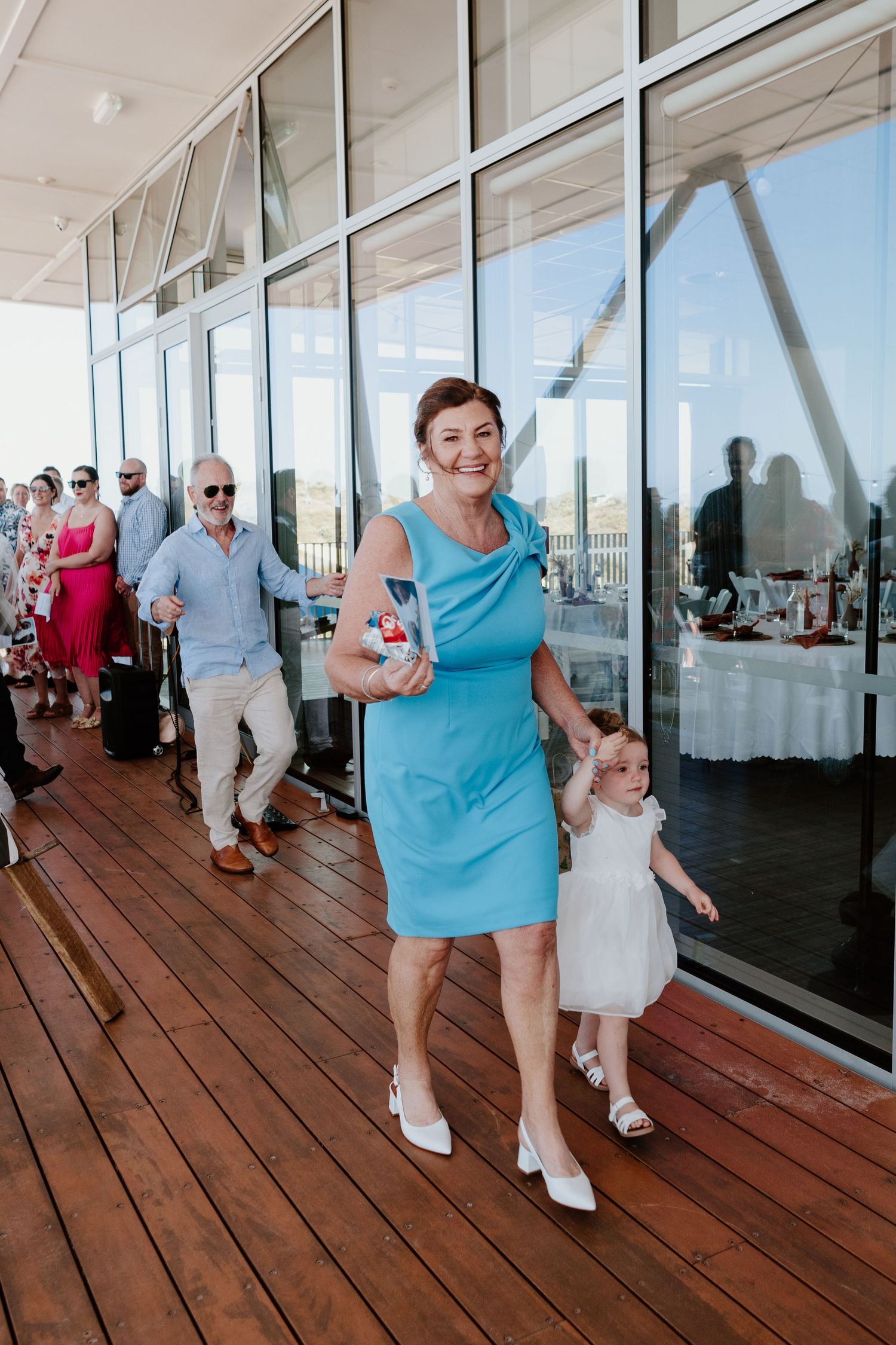 Wedding guests in bright attire walk and dance along a covered outdoor deck. Papamoa Surf Lifesaving Club Wedding