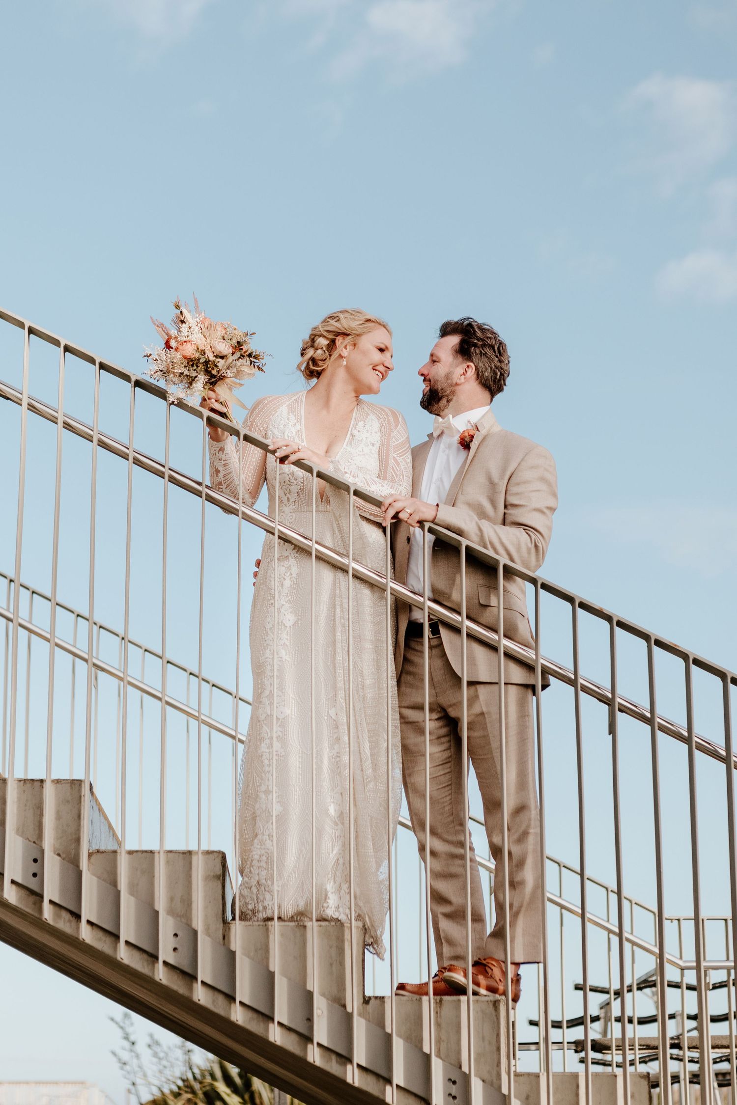 A wedding couple stands together on an outdoor staircase against a blue sky backdrop holding a bouquet.