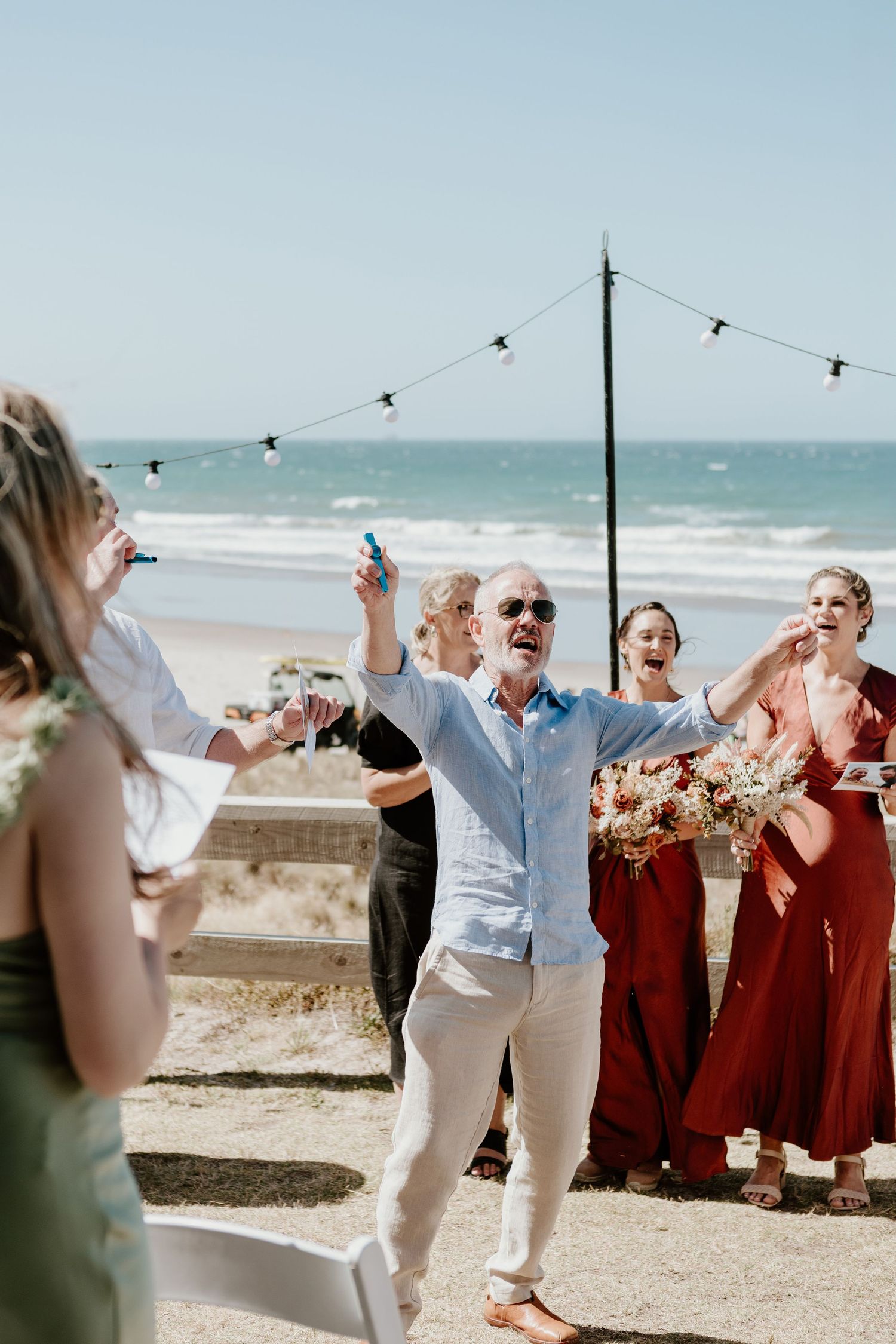 A wedding celebration at a beach with people laughing and dancing on a sunny day with string lights overhead.