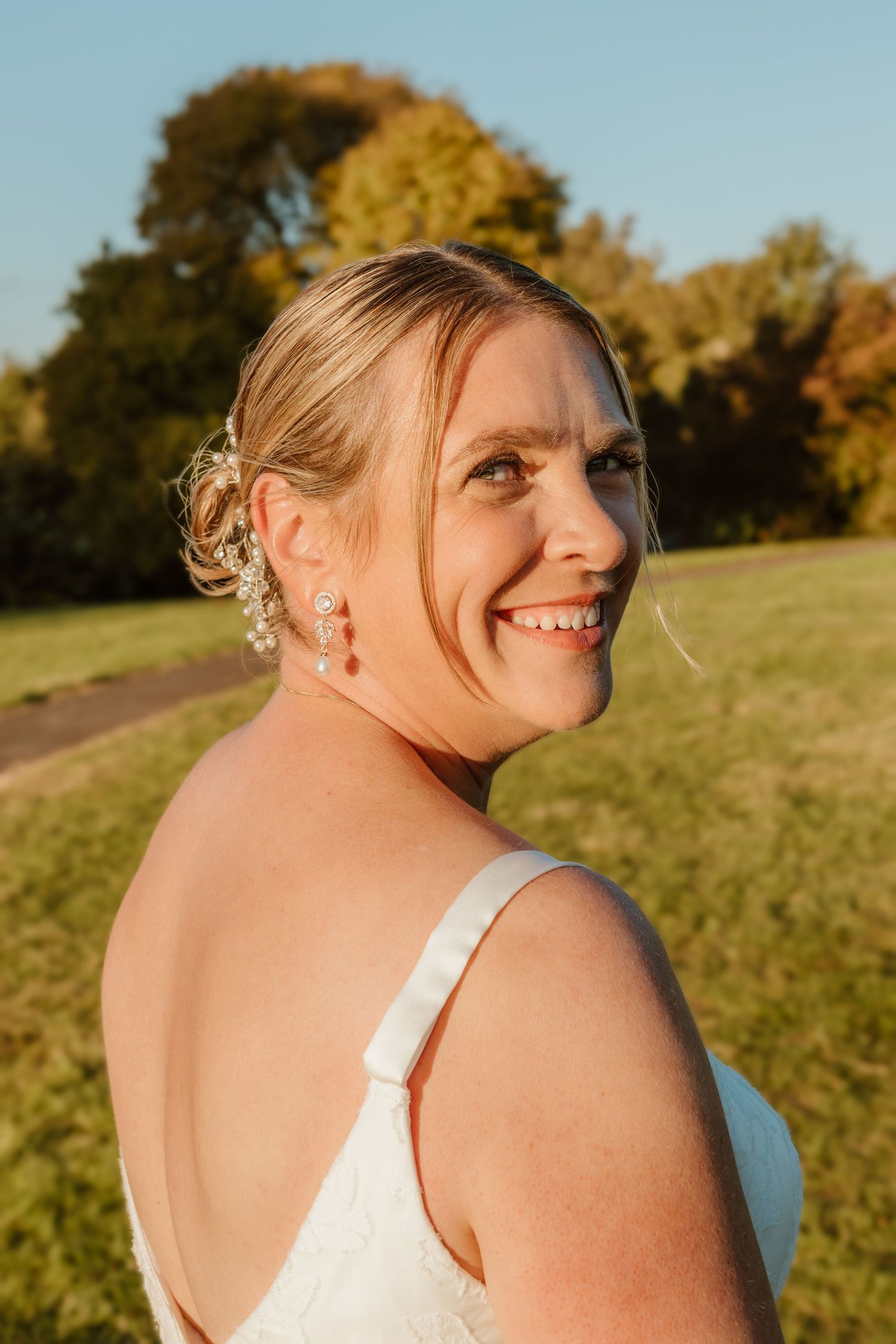 A bride wearing a white dress turns to smile over her shoulder in a sunlit outdoor setting with autumn trees in background.