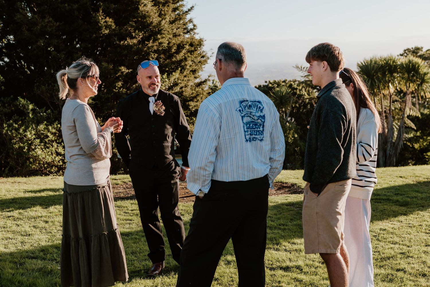 Group of people stand together outdoors on a grassy hillside during golden hour with evergreen trees in the background.