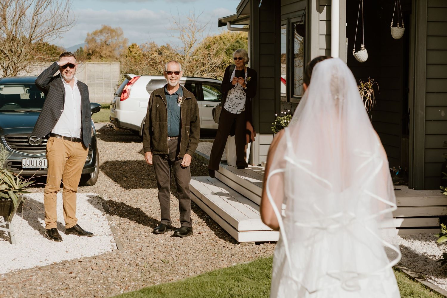 Wedding party members react with joy as the bride approaches on the porch of a rustic venue on a sunny day.