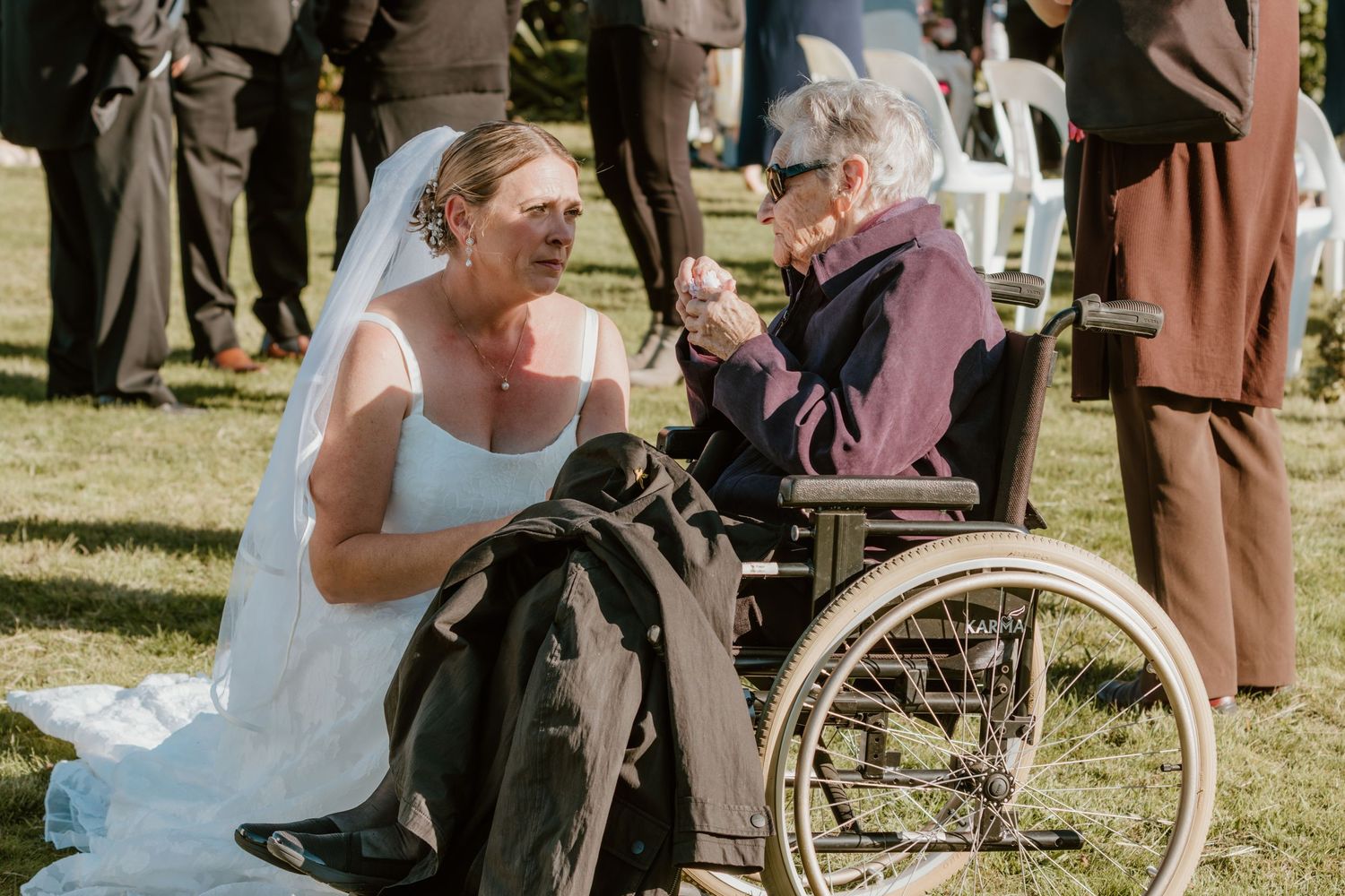 A bride in a white wedding dress kneels beside an elderly person in a wheelchair during an outdoor ceremony.