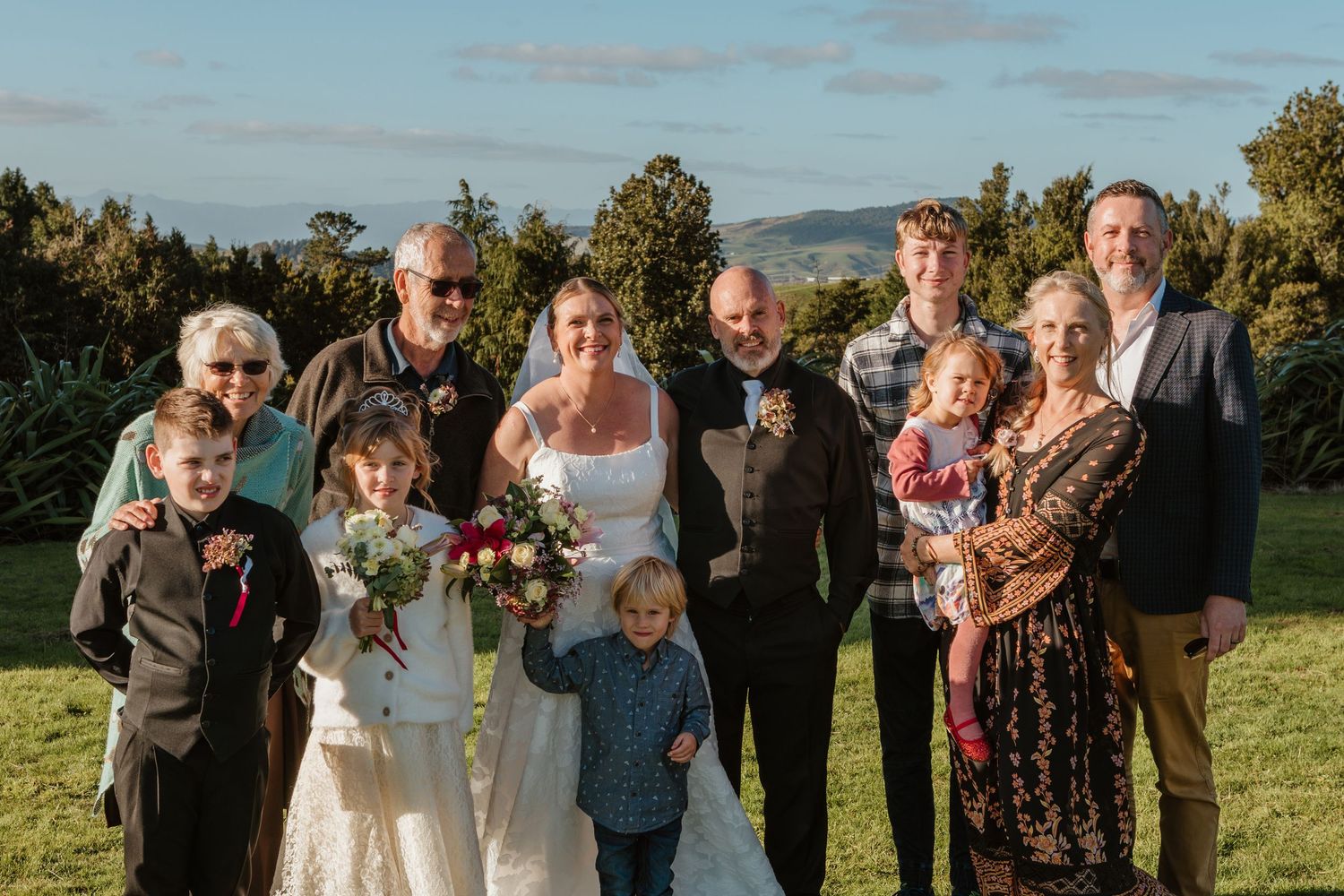 Wedding party poses together on a grassy hillside during golden hour sunset celebration with mountains in background.