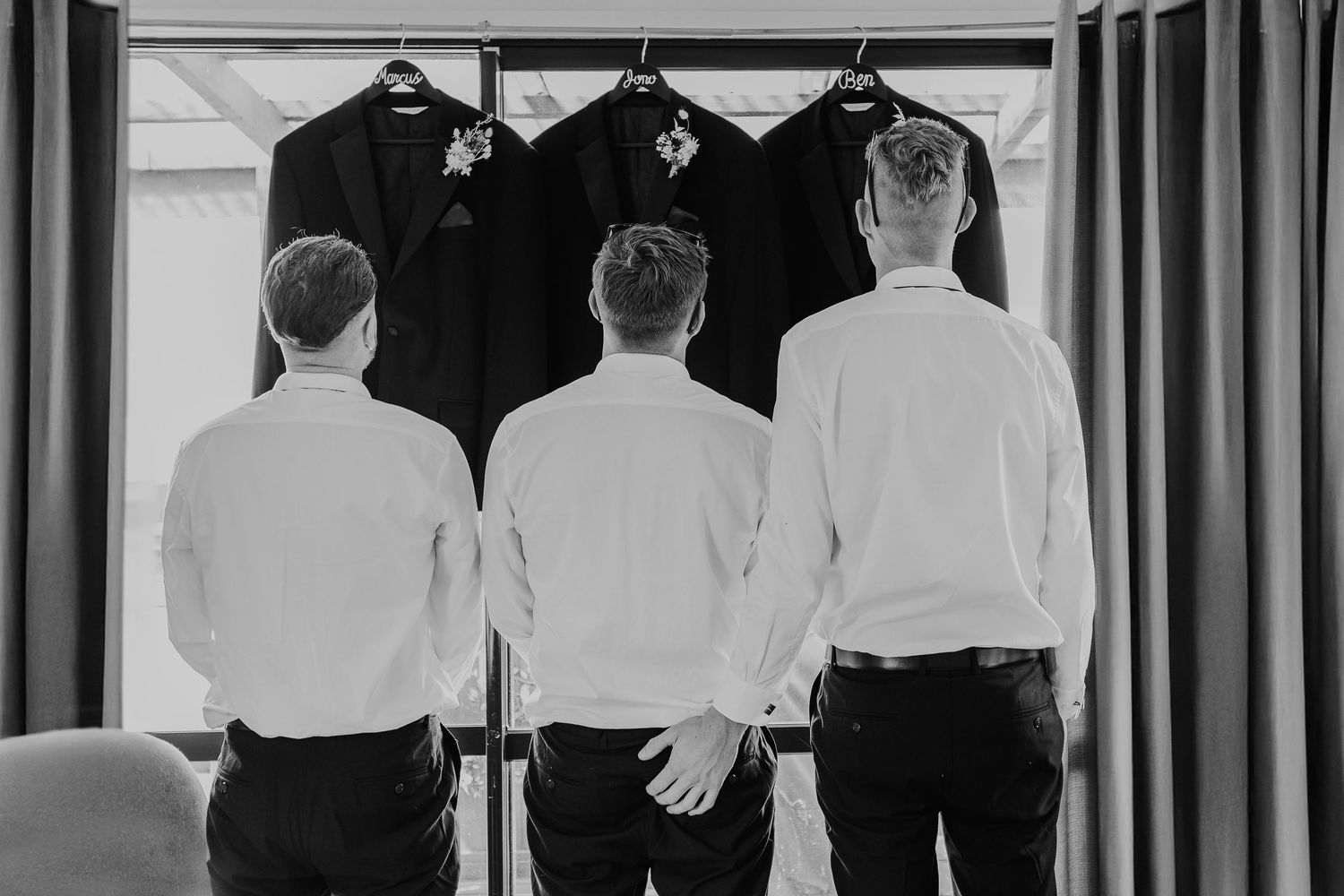 Black and white photo of groomsmen getting ready, viewed from behind as they put on formal attire and jackets.