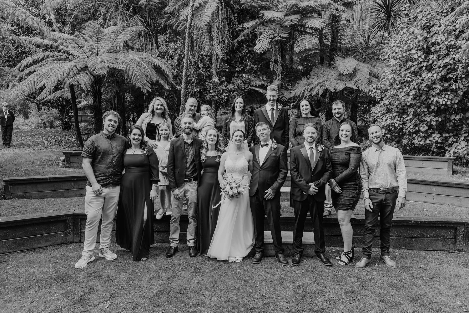 A wedding party stands together in formal attire for a black and white photograph in a lush garden setting.