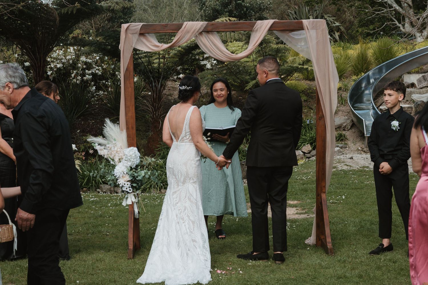 Wedding ceremony guests gather under white draped canopy in outdoor garden setting during sunset.