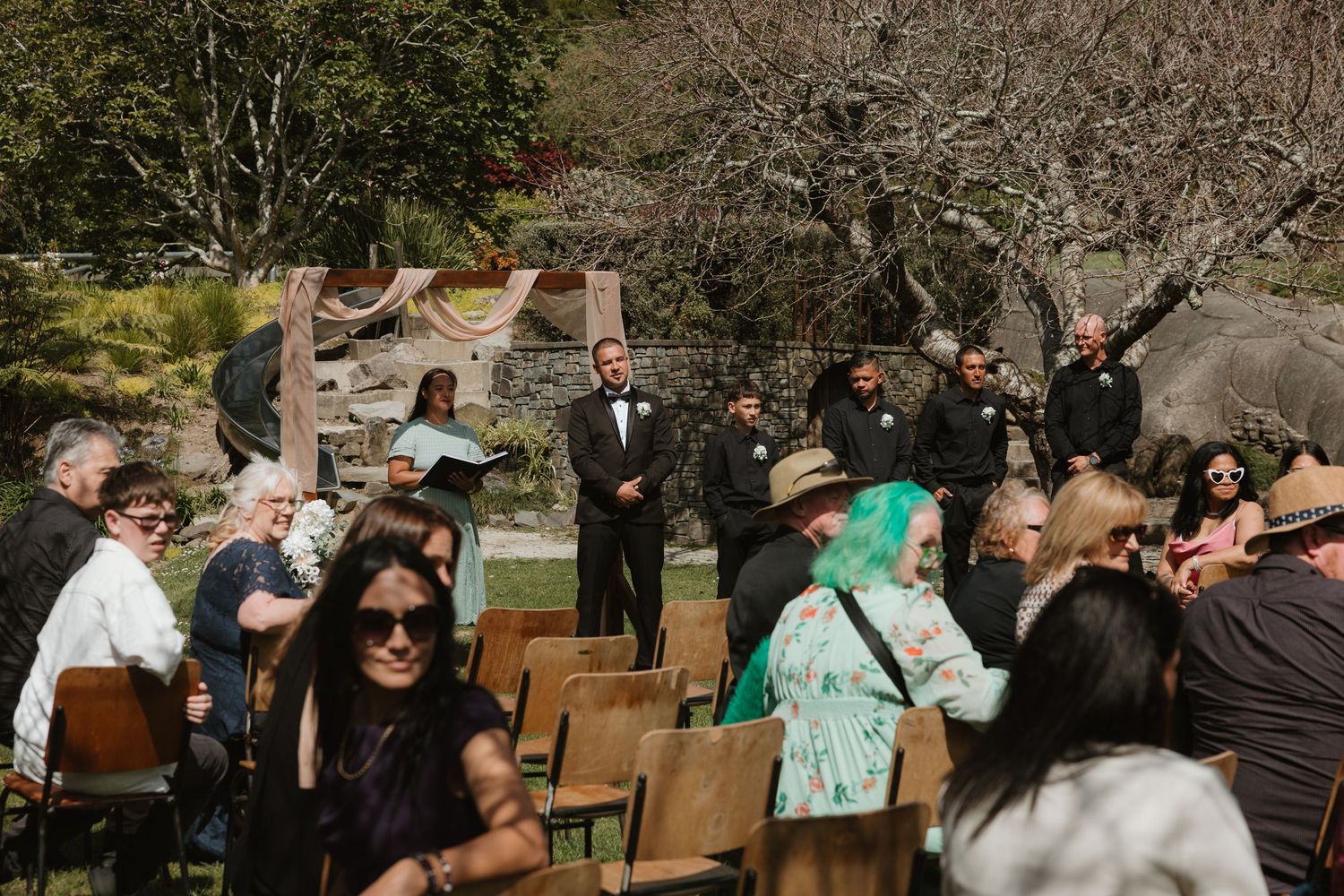 A group of people enjoying an outdoor celebration in a natural setting with wooden chairs and trees in the background.
