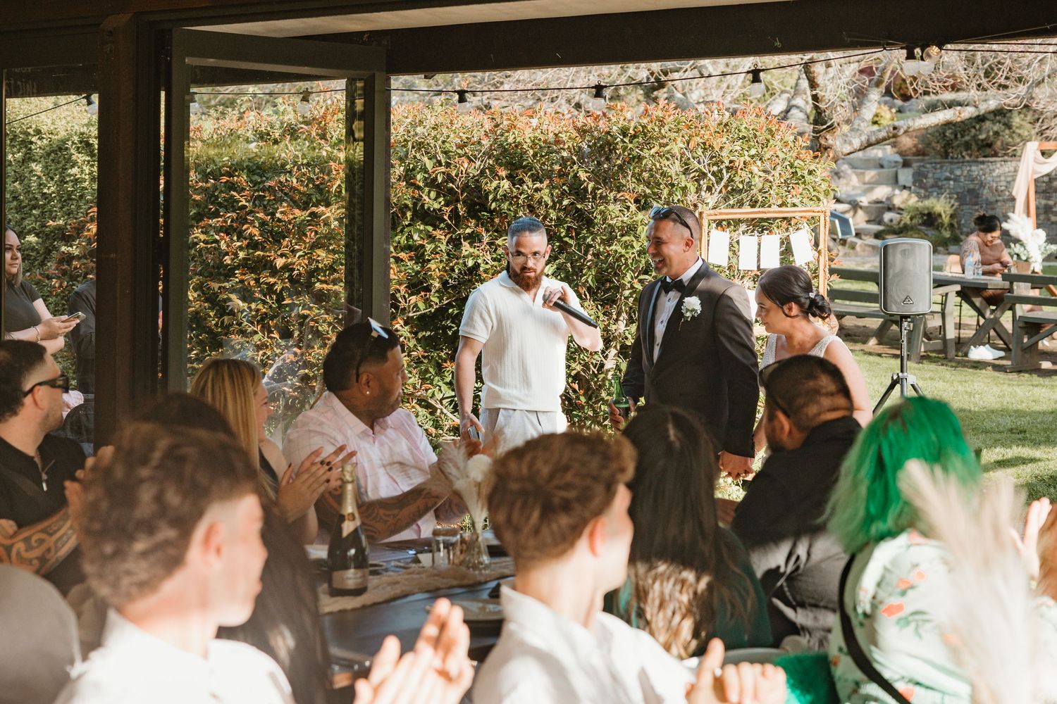 Wedding guests gather under a covered outdoor patio area during a daytime celebration in a garden setting.