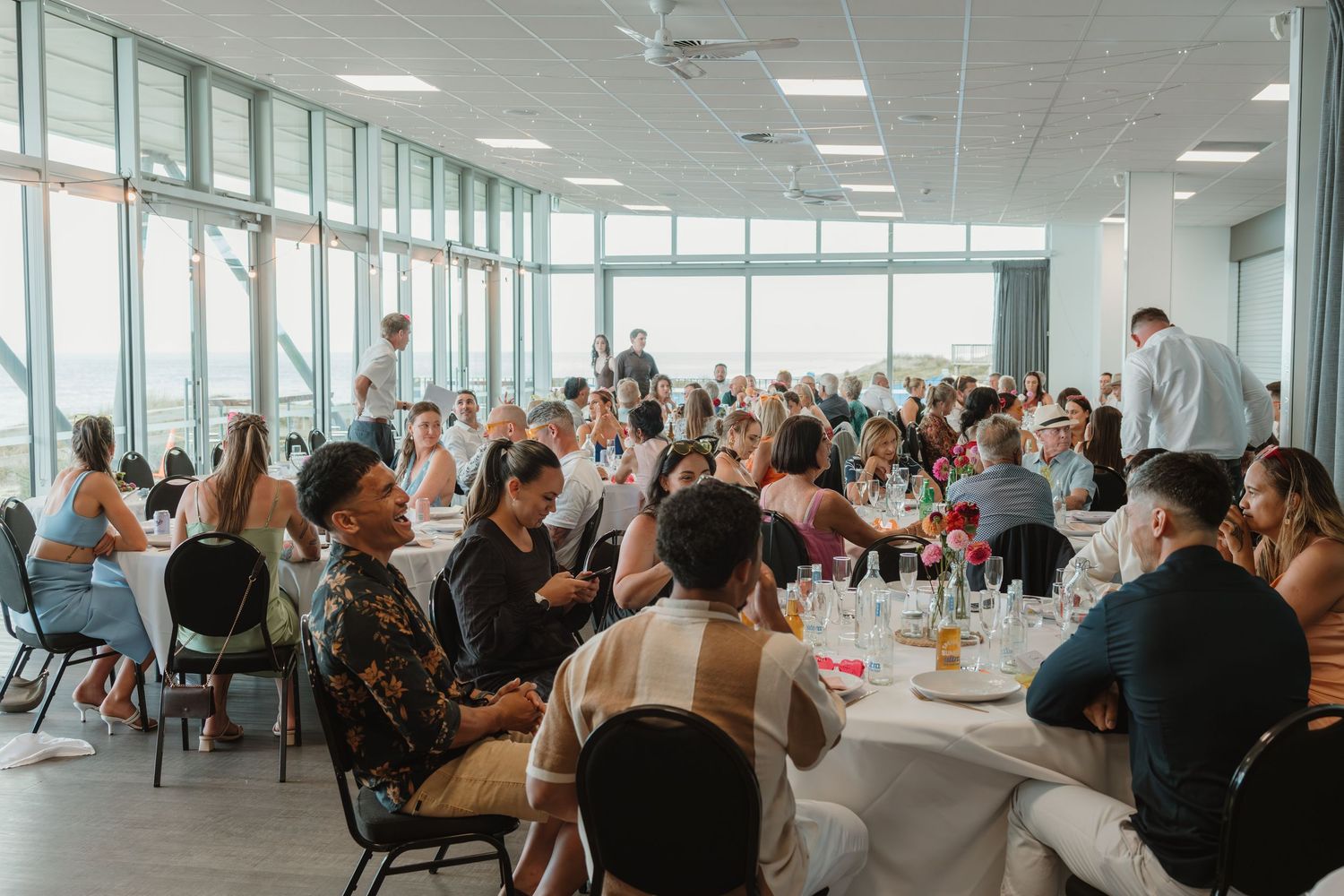 Elegant wedding reception banquet hall filled with guests seated at round tables near large windows.