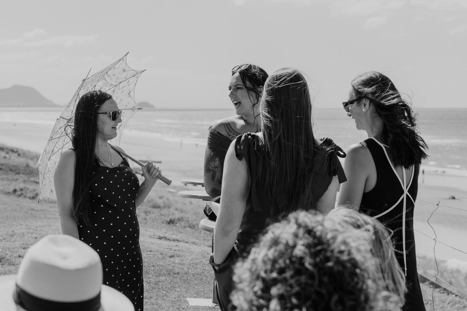 Women in summer dresses chat on a scenic coastal overlook, one holding a lace parasol, in black and white photography.
