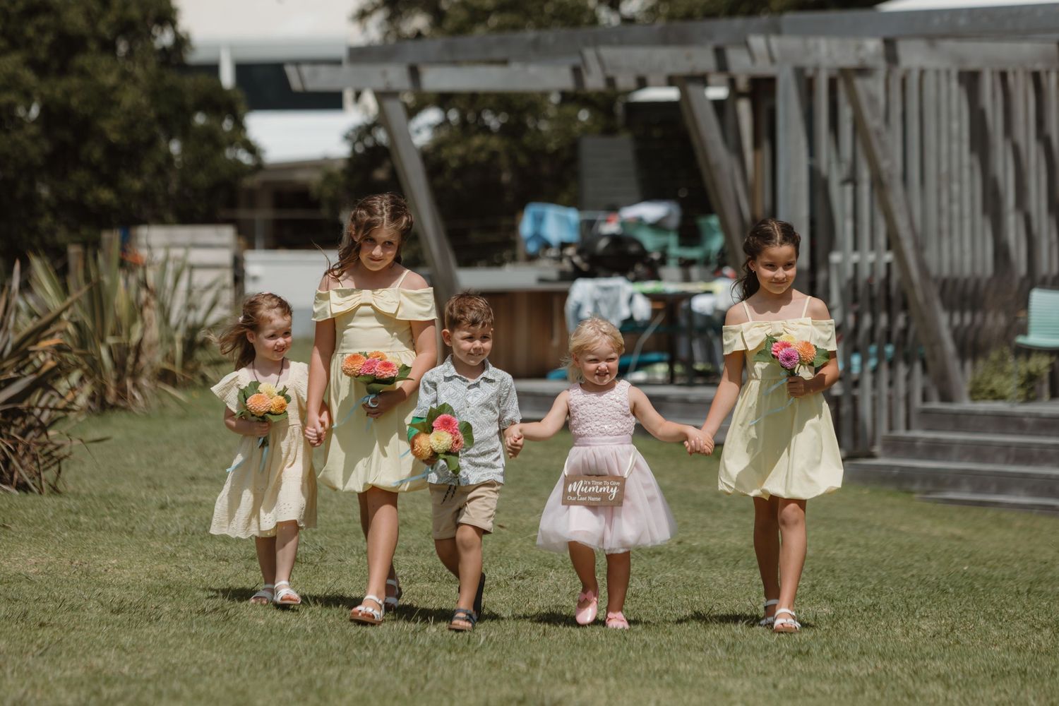 Five children in pastel outfits walk together on green grass, holding bouquets, dressed as flower girls and ring bearer.