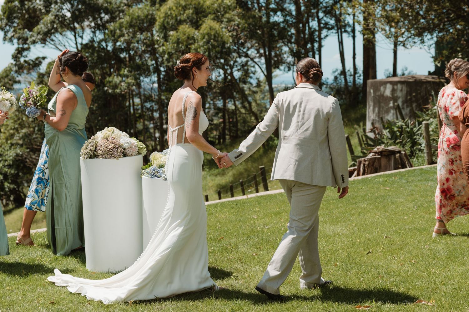 Bride and groom walk hand in hand at an outdoor wedding ceremony on a sunny day with lush green lawn.