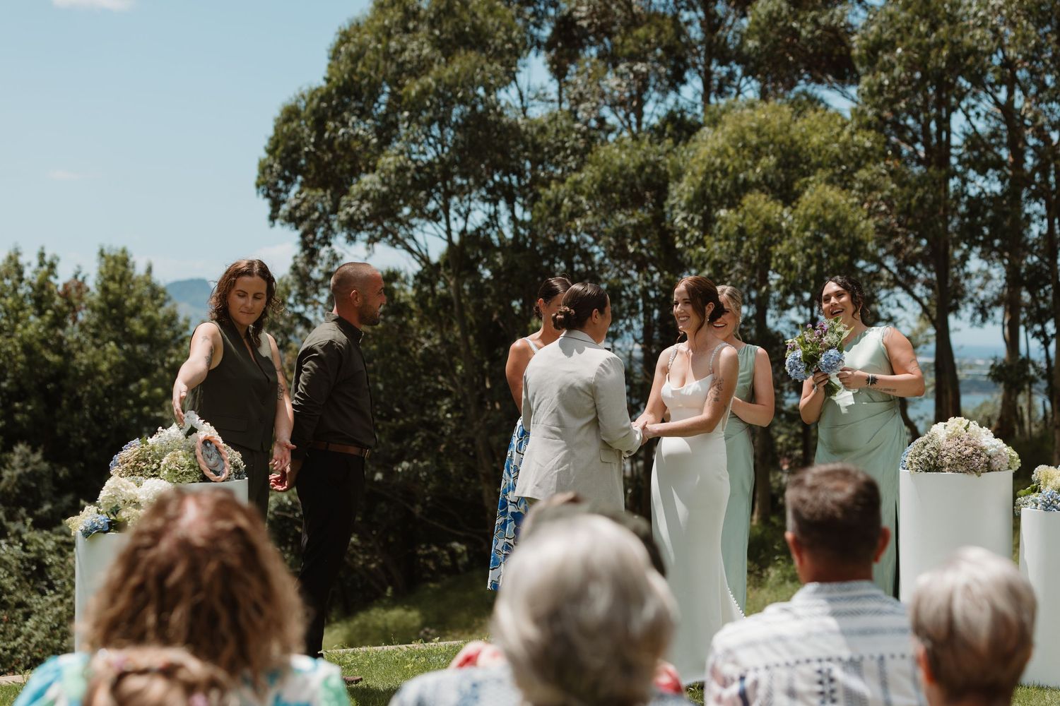 Outdoor wedding ceremony with bridesmaids in sage green dresses and guests seated on a scenic hilltop.