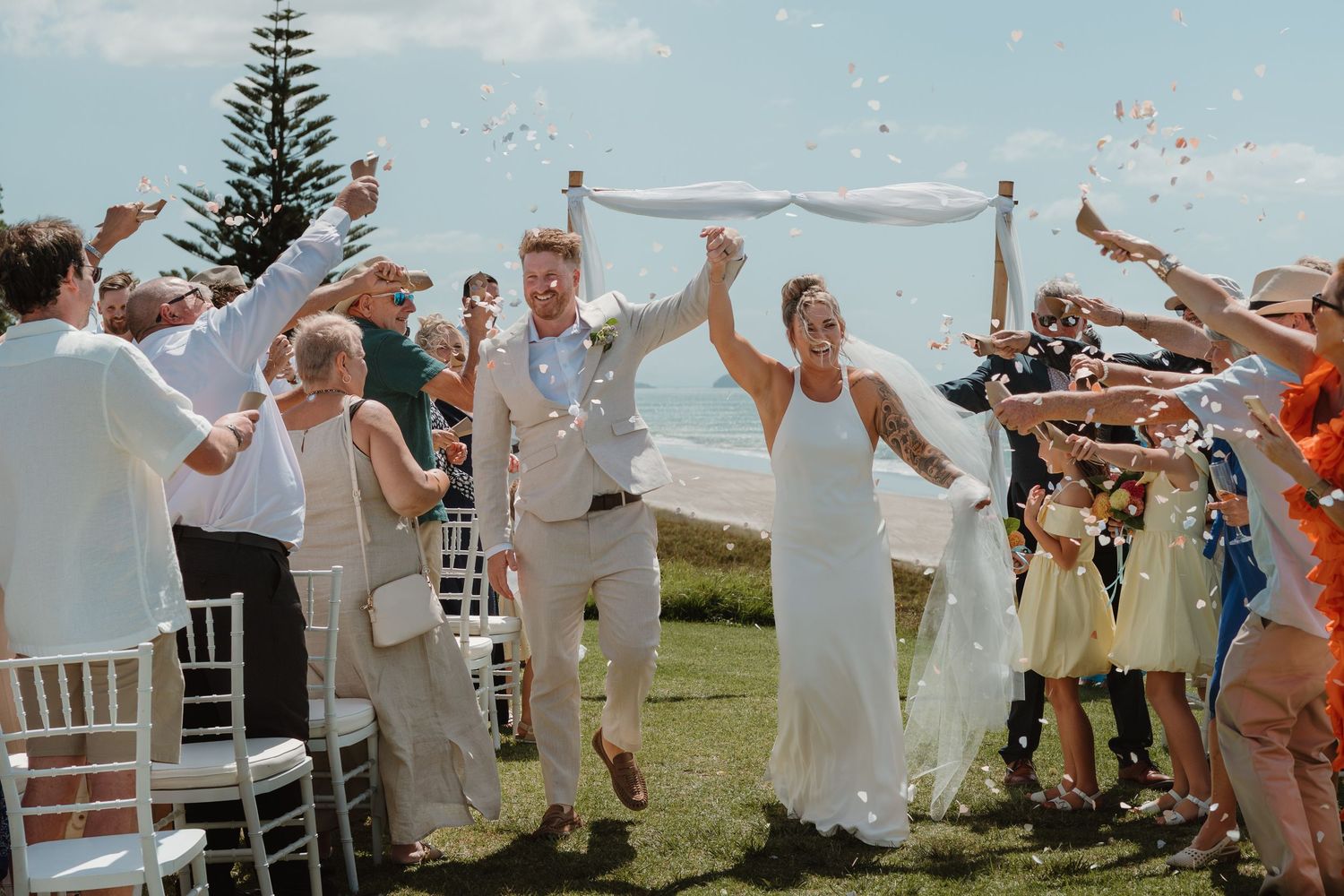 Joyful newlyweds walk down the aisle as guests toss confetti at an outdoor beach wedding ceremony.