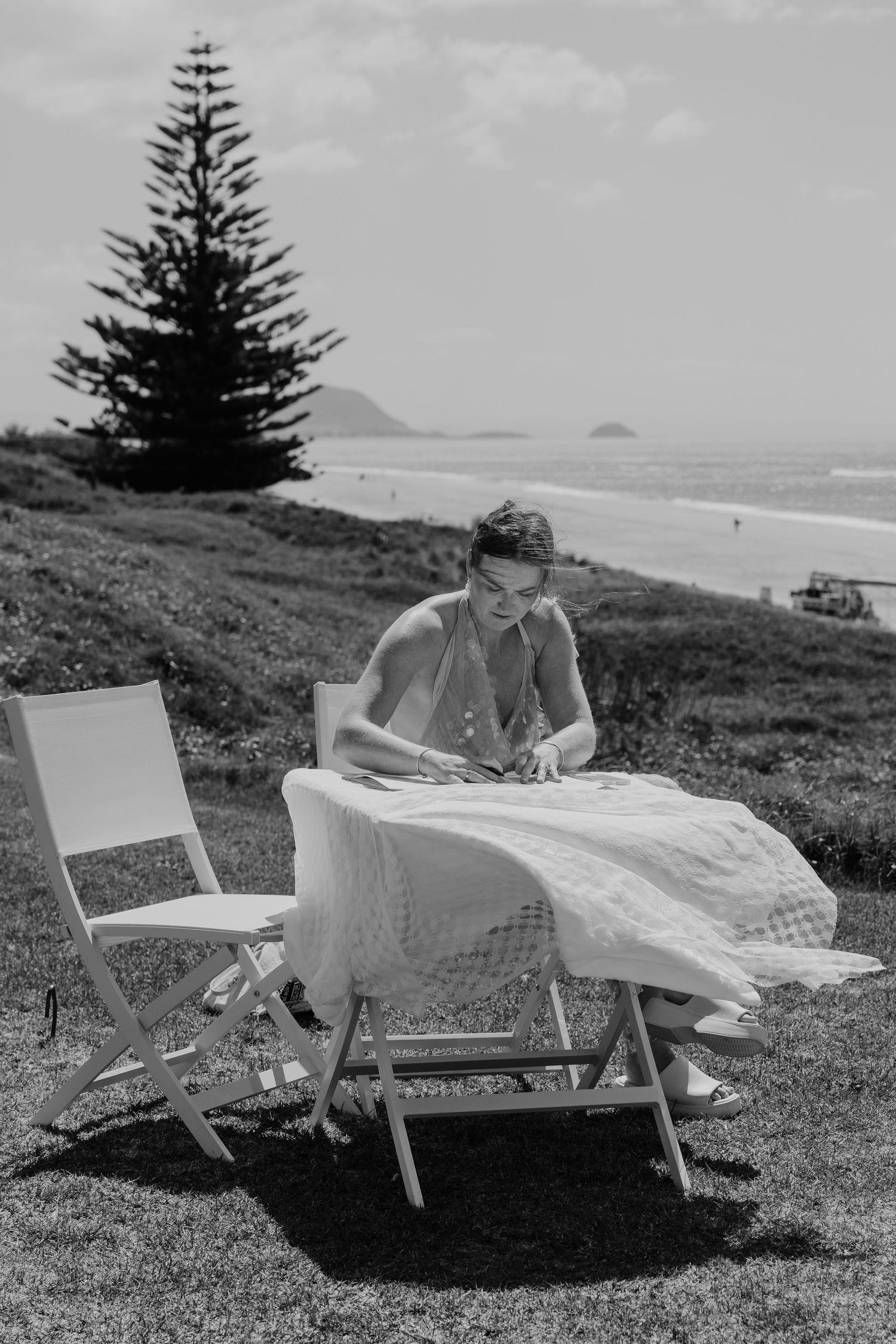 Black and white photo of a woman arranging fabric on a chair outdoors near a beach with a Norfolk pine tree.