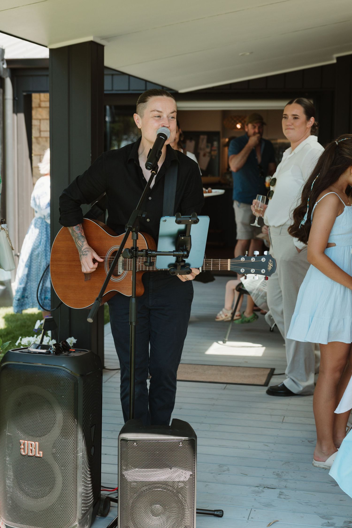 A musician in black plays acoustic guitar and sings into a microphone at an outdoor wedding reception with guests watching.