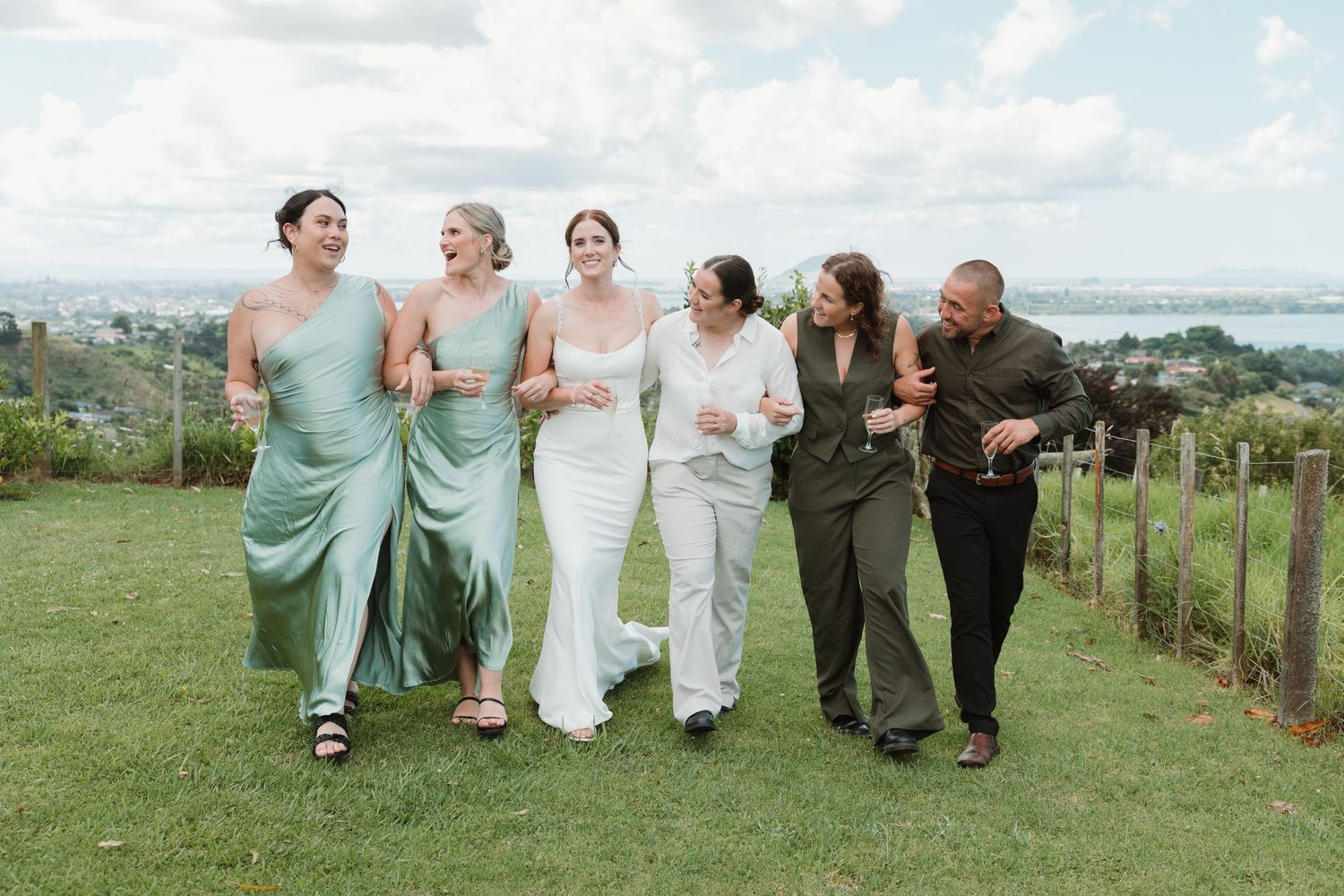 Bride and bridesmaids in sage green dresses walking on a grassy hillside with panoramic views on a wedding day.