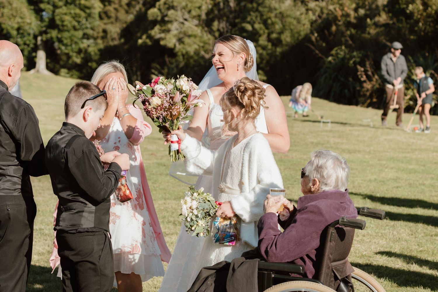 Wedding guests gather on a sunny lawn during an outdoor ceremony holding bouquets of flowers and drinks.