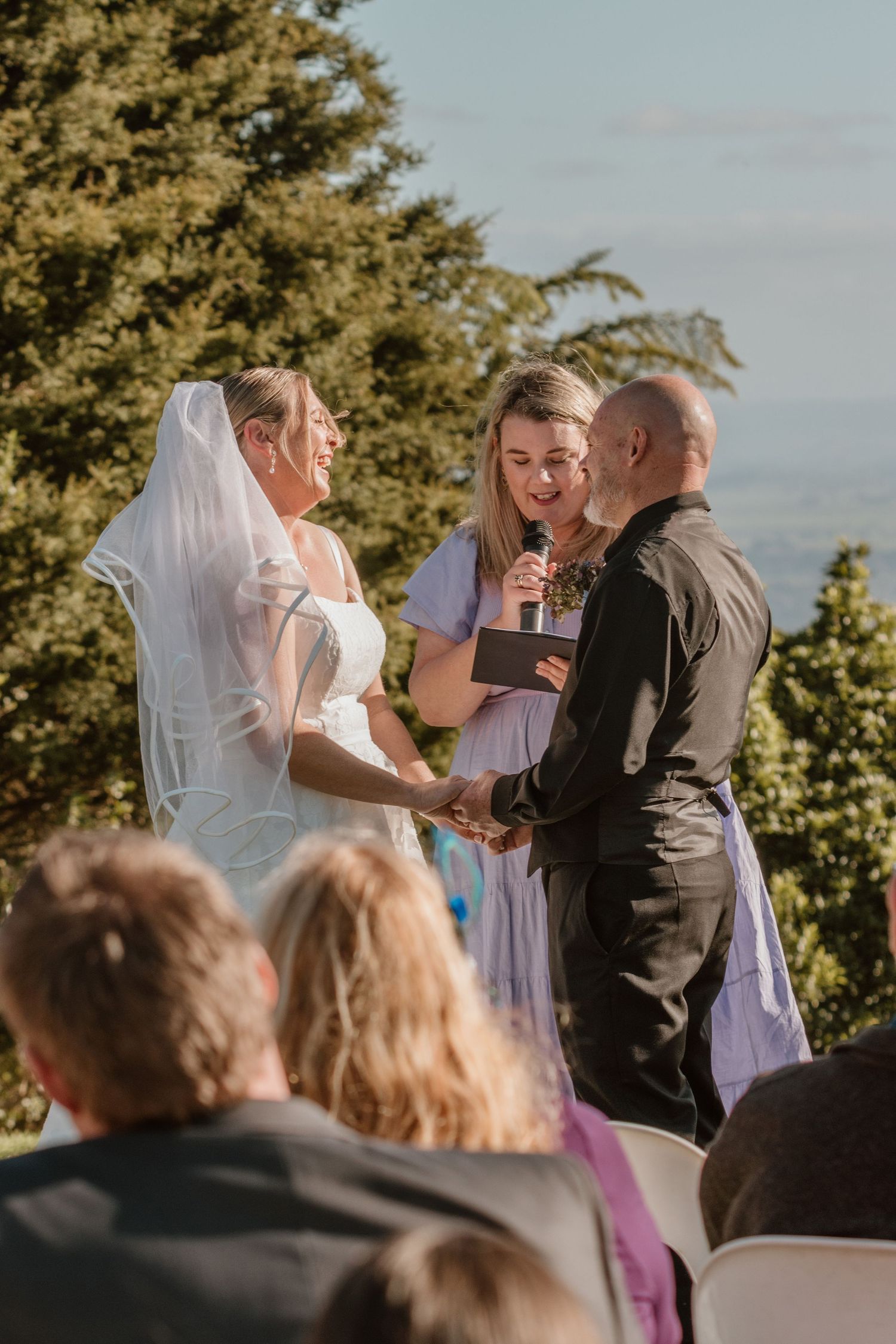 An outdoor wedding ceremony takes place on a hilltop with evergreen trees and guests seated in the background.