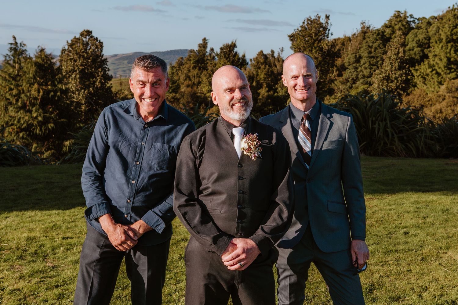 Three people in formal attire stand together smiling on a grassy hillside with autumn trees in the background.