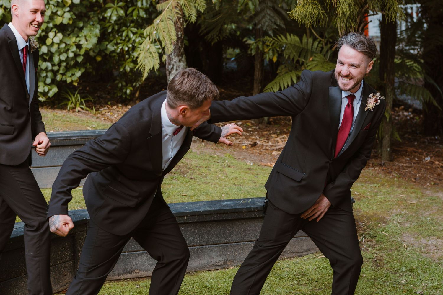 Men in black suits playfully pose and laugh together outdoors in a garden setting.