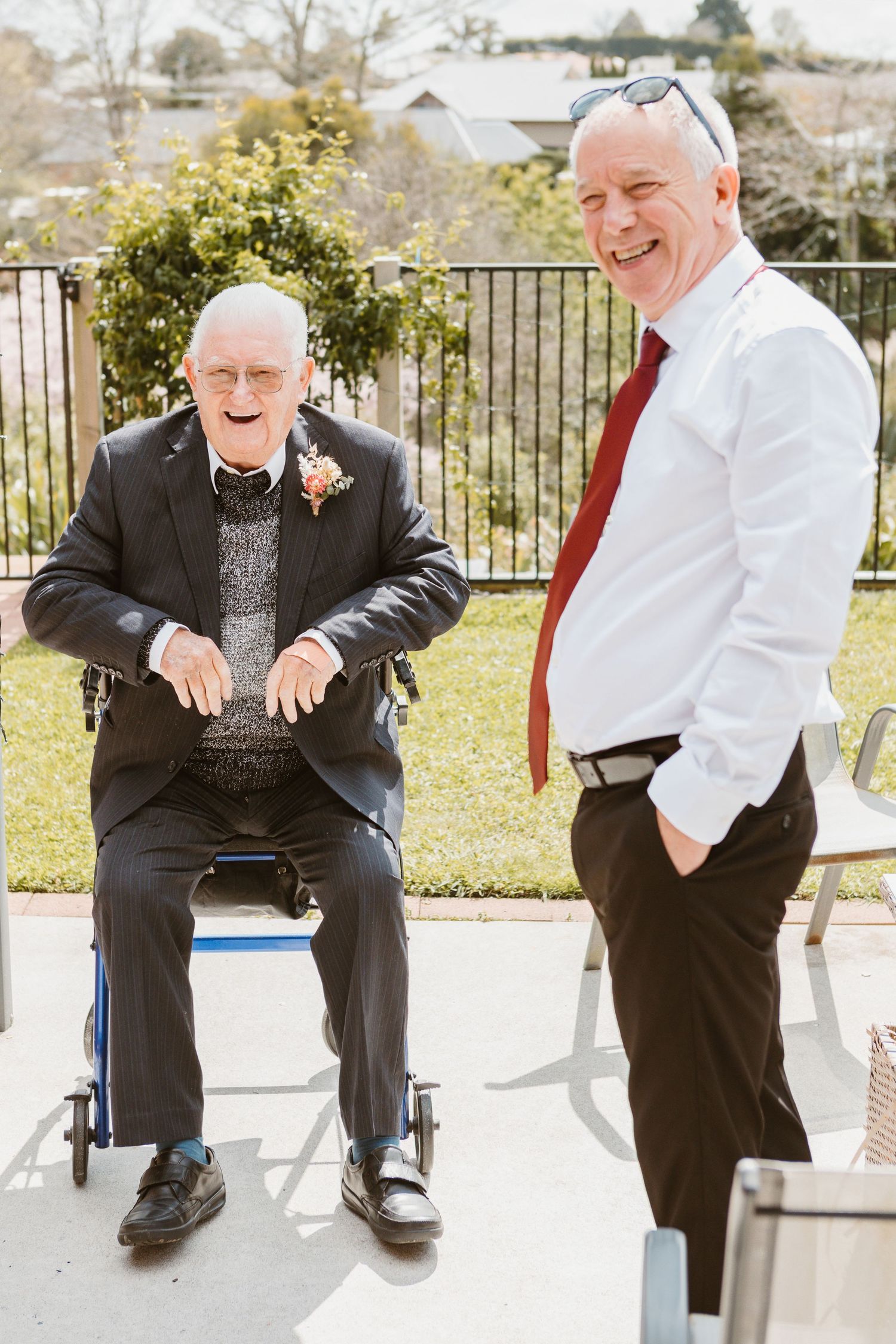 Two people laughing and enjoying a sunny moment together on an outdoor patio with garden scenery in the background.