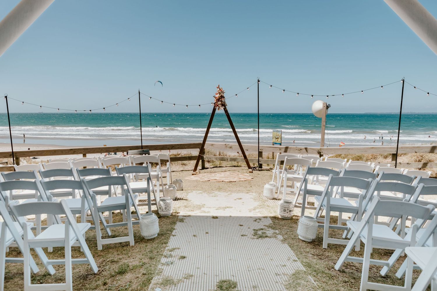 Beach wedding ceremony setup with white chairs and wooden arch decorated with string lights overlooking the ocean.