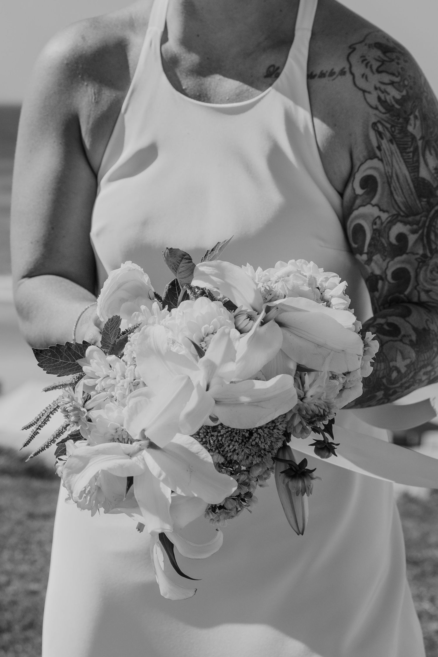 Black and white photo of a bride in white dress holding a floral bouquet beside a tattooed groom at a wedding.