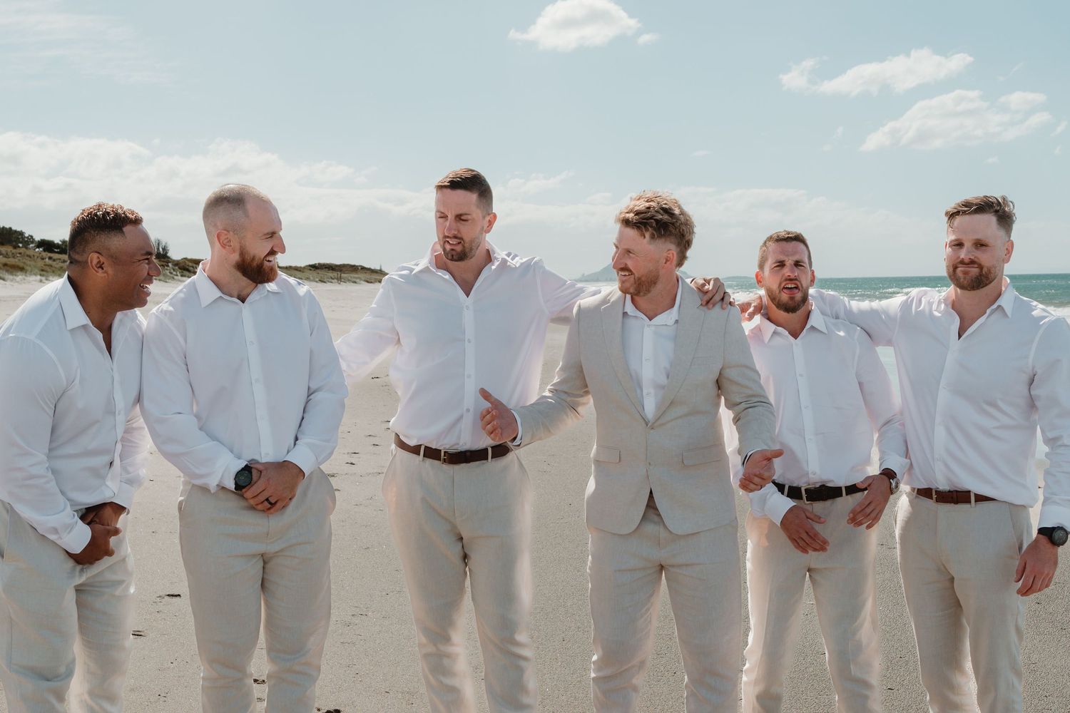 Six groomsmen in white shirts and khaki pants laughing together on a sunny beach wedding day.